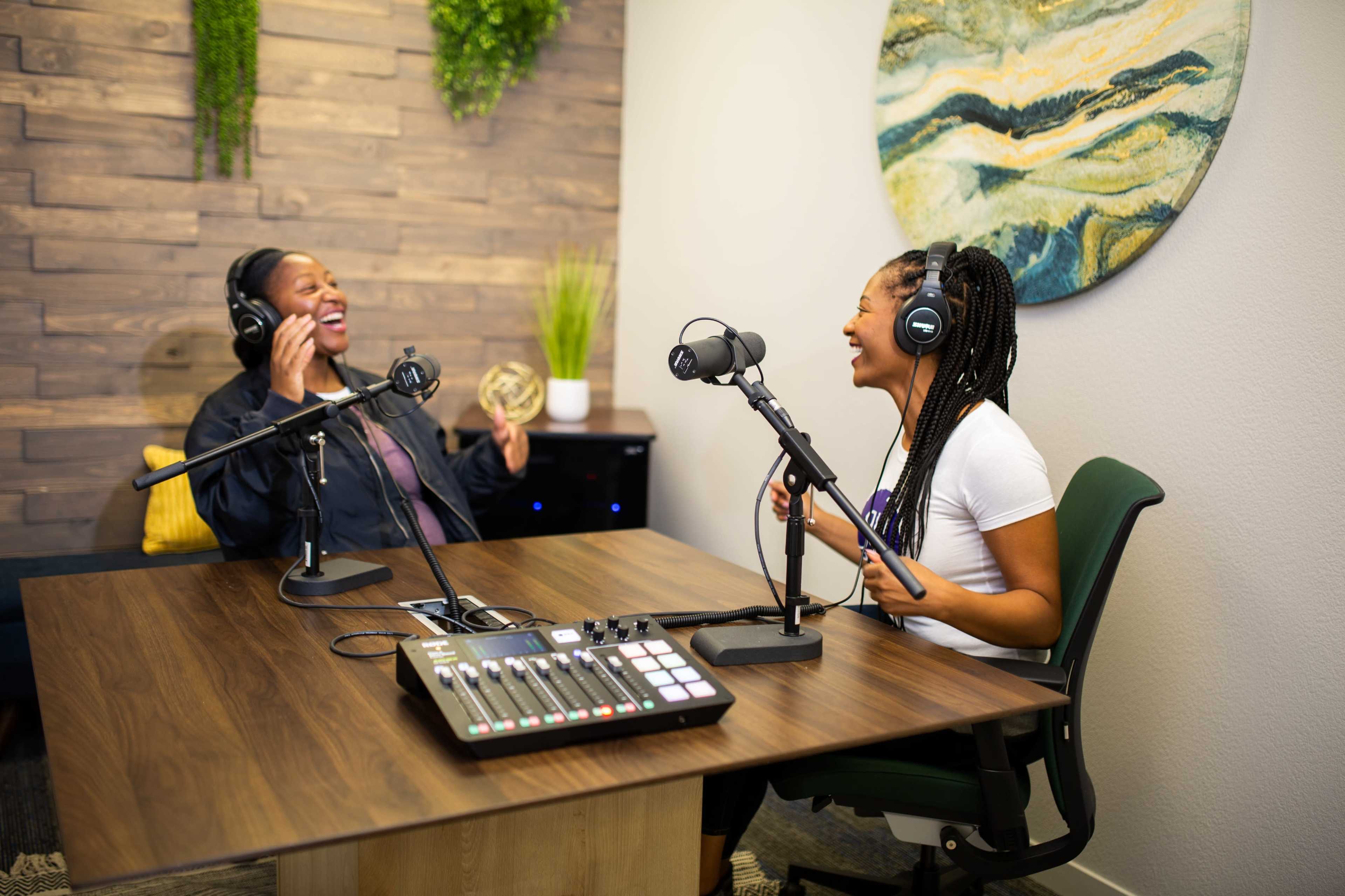 Two women are seated at a table with microphones, laughing and engaging in a conversation in a recording studio.
