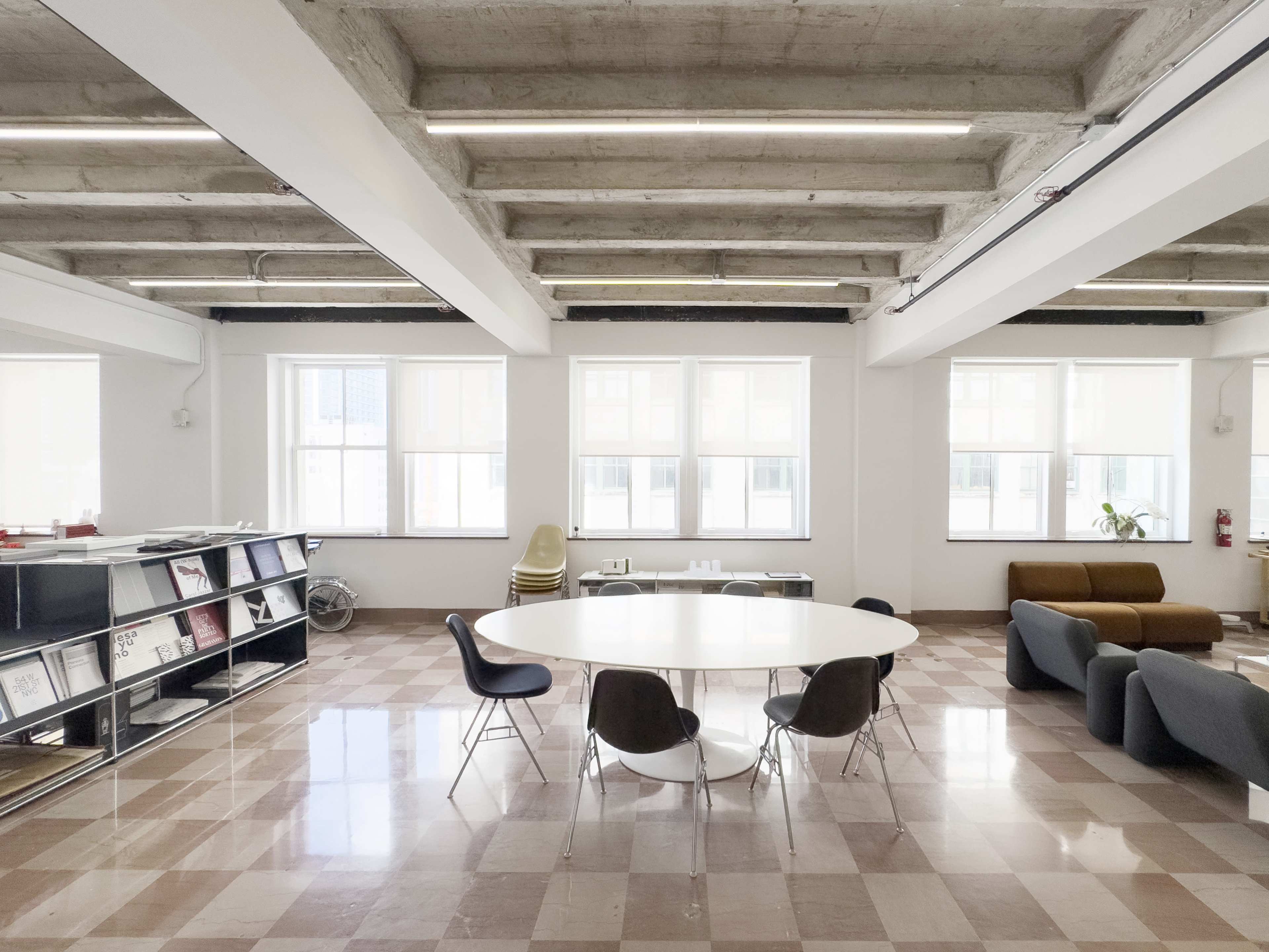 The image shows a modern office space with a round table surrounded by chairs, large windows allowing natural light, and a seating area with a sofa.