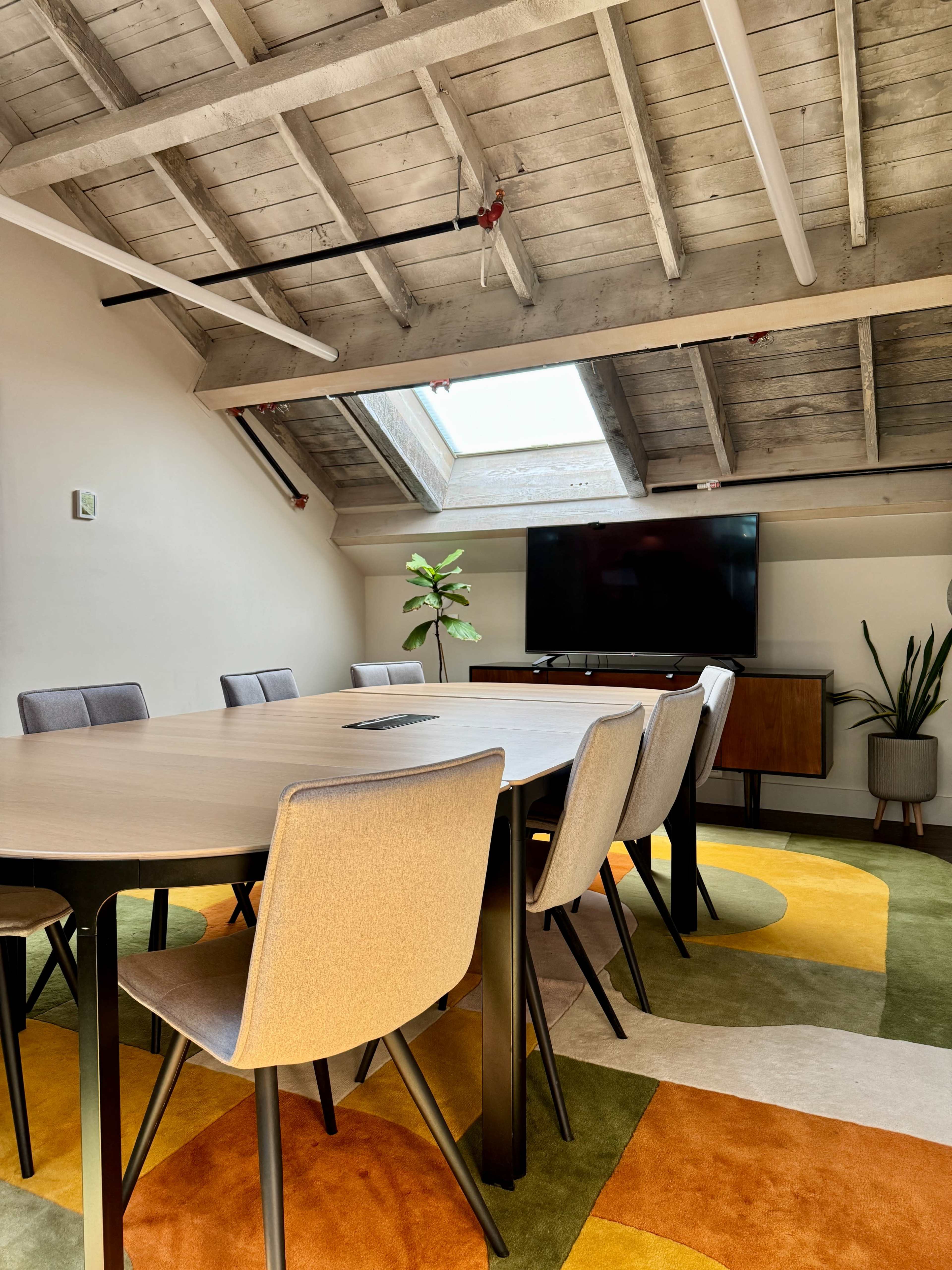 A brightly lit conference room features a large round table surrounded by gray chairs, with a television mounted on the wall and a colorful patterned rug on the floor.