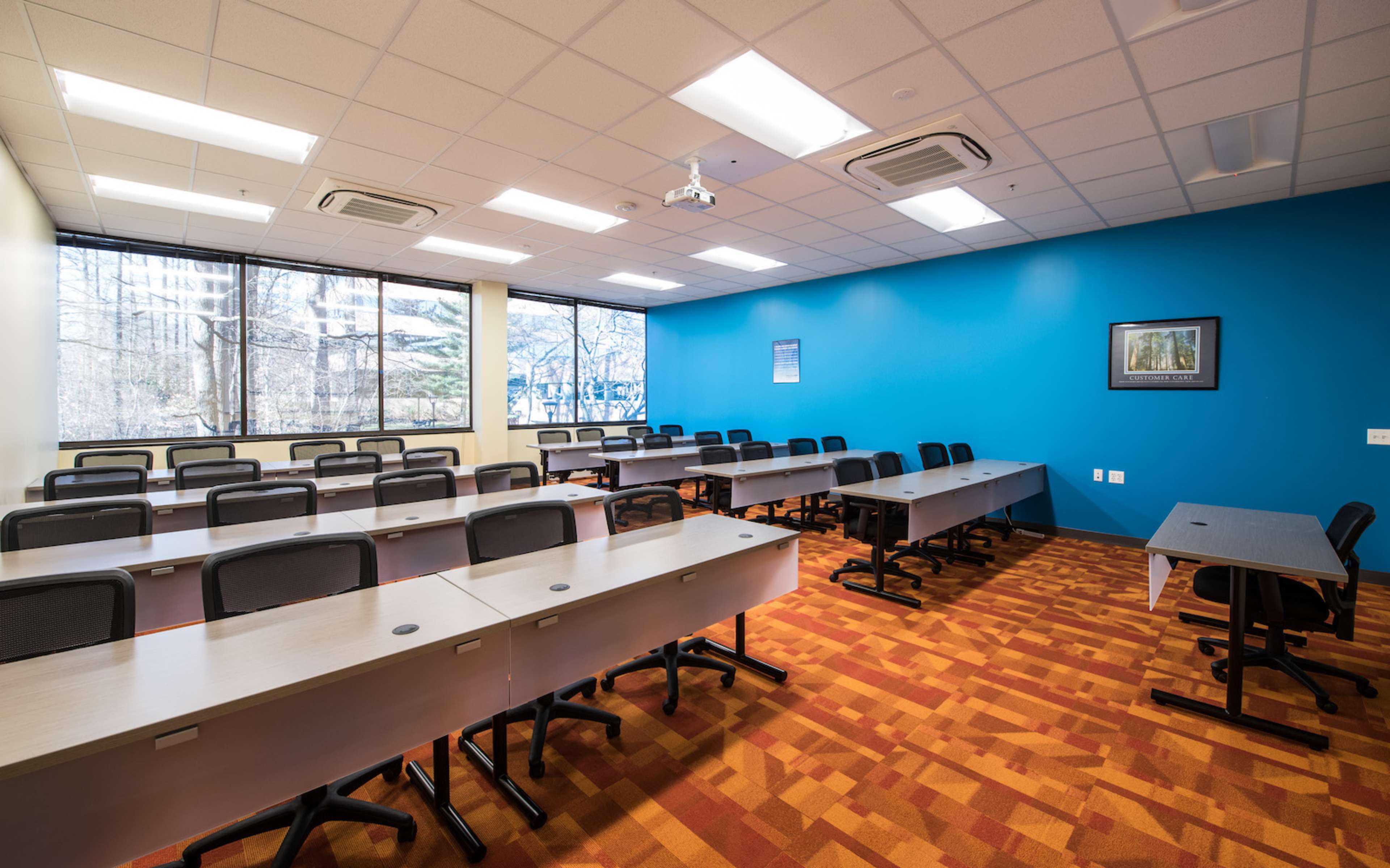 The image shows a modern classroom with rows of tables and chairs, large windows overlooking trees, and one blue accent wall.