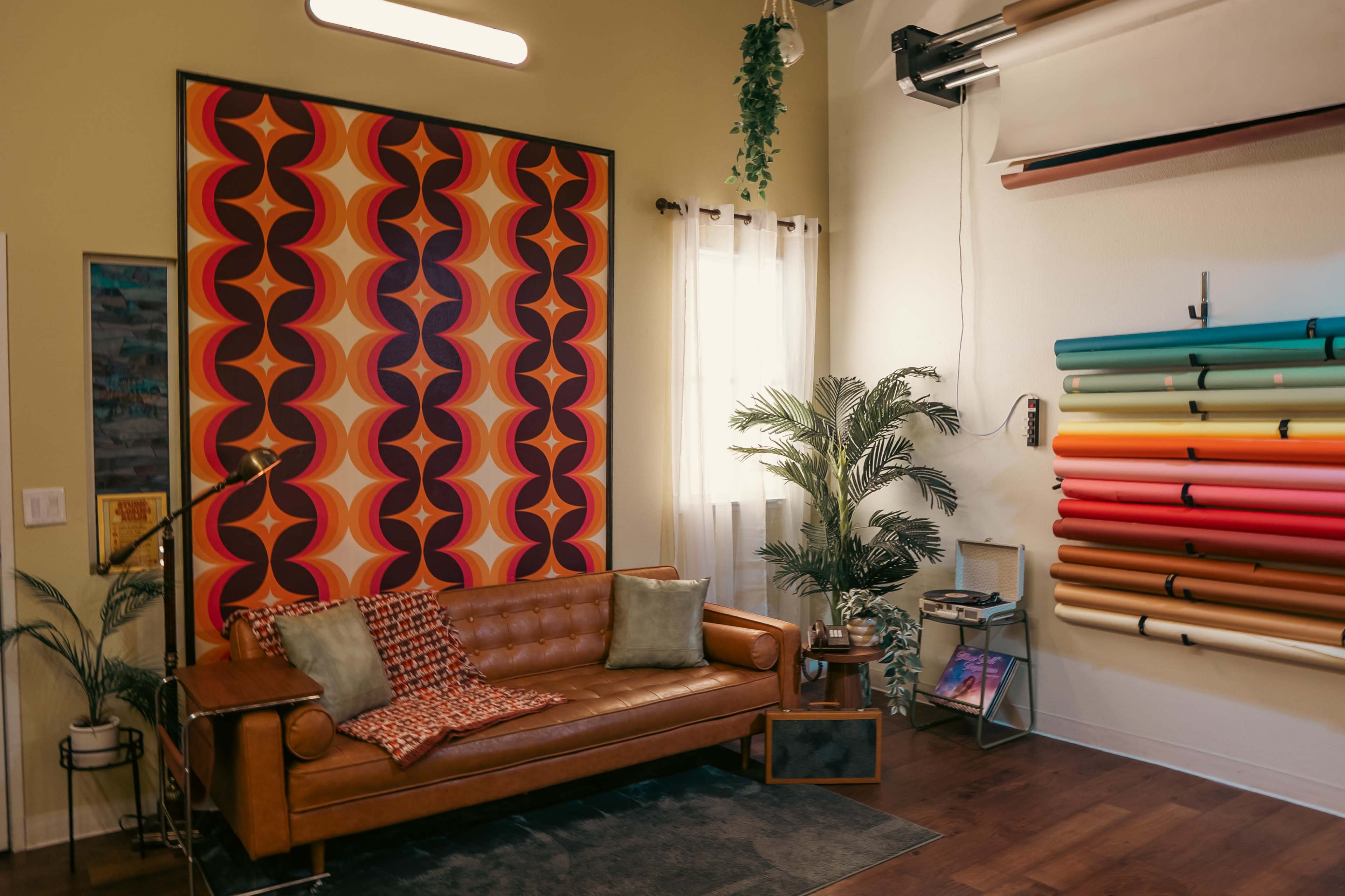 The image shows a stylish living room featuring a brown couch with patterned cushions, colorful rolls of paper on the wall, and large windows with sheer curtains.