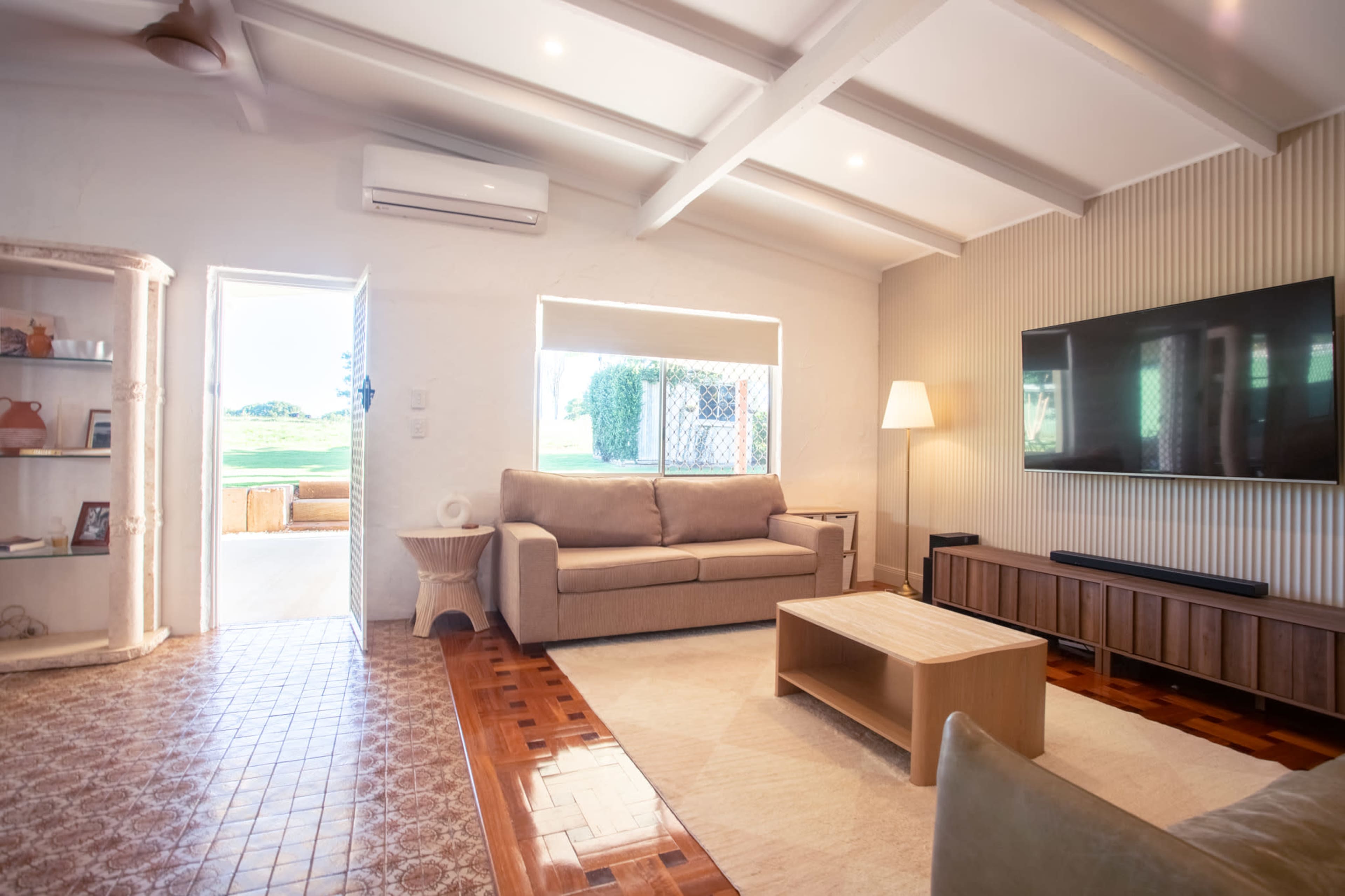 The room features a modern living area with a large sofa, a coffee table, and a television mounted on the wall, illuminated by natural light coming from a nearby window.