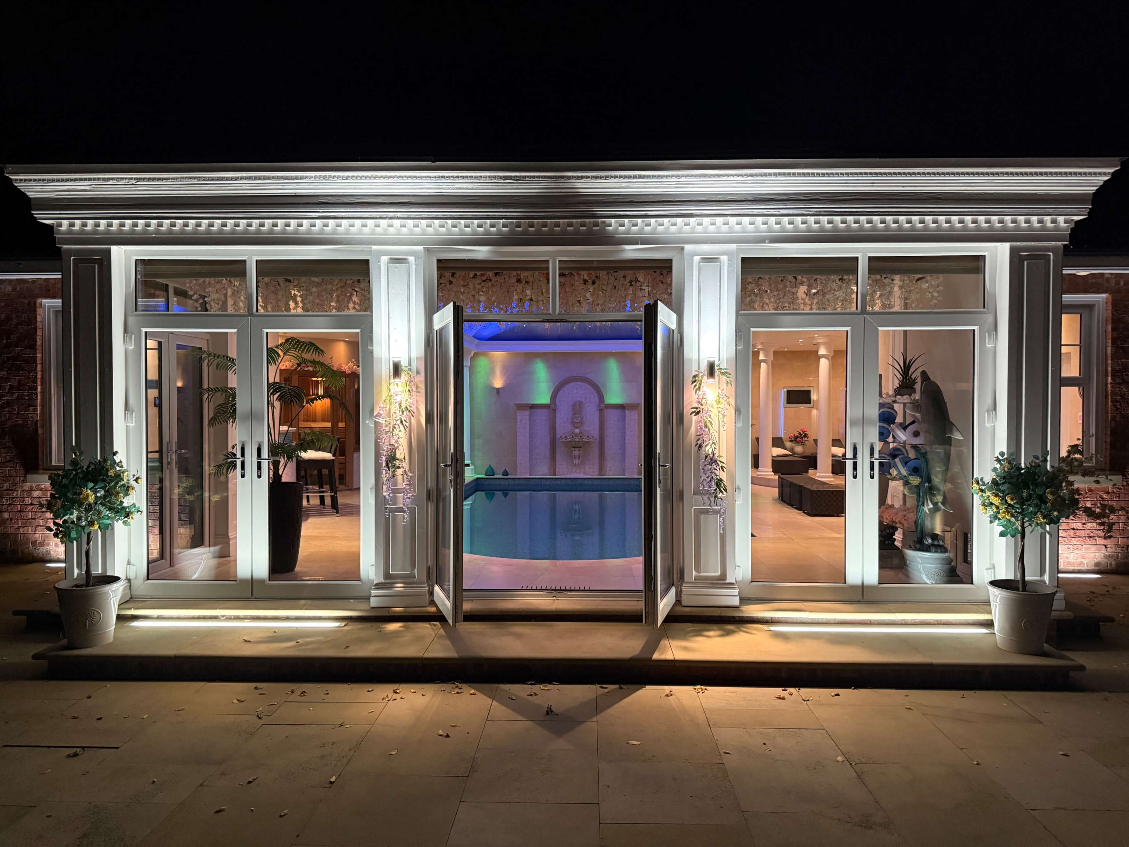 A modern building features large open doors leading to an illuminated indoor pool area, with decorative plants on either side.