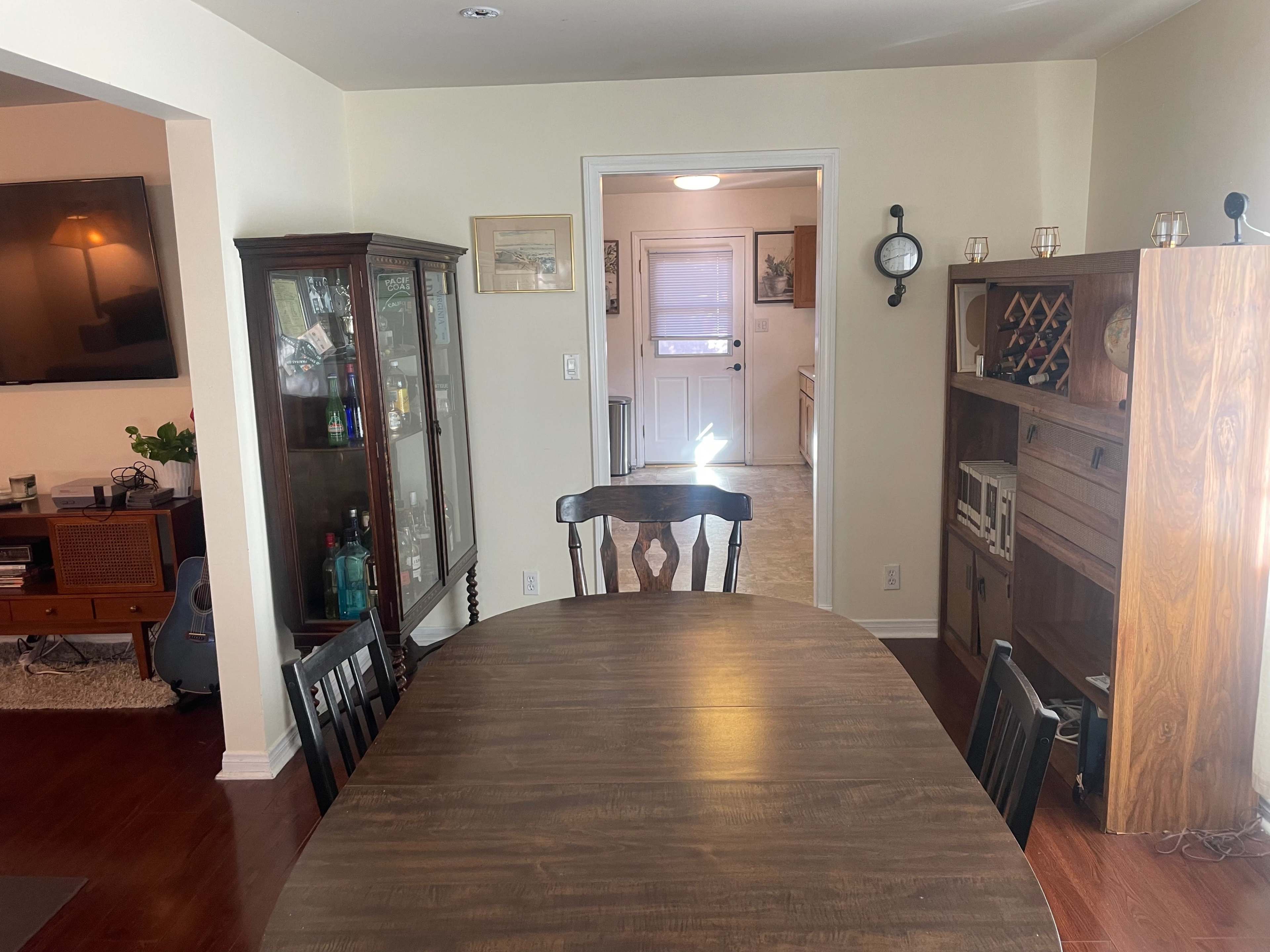 A dining area with a brown wooden table and chairs, and wooden cabinets displaying glassware, leading to a doorway with a light-colored wall in the background.