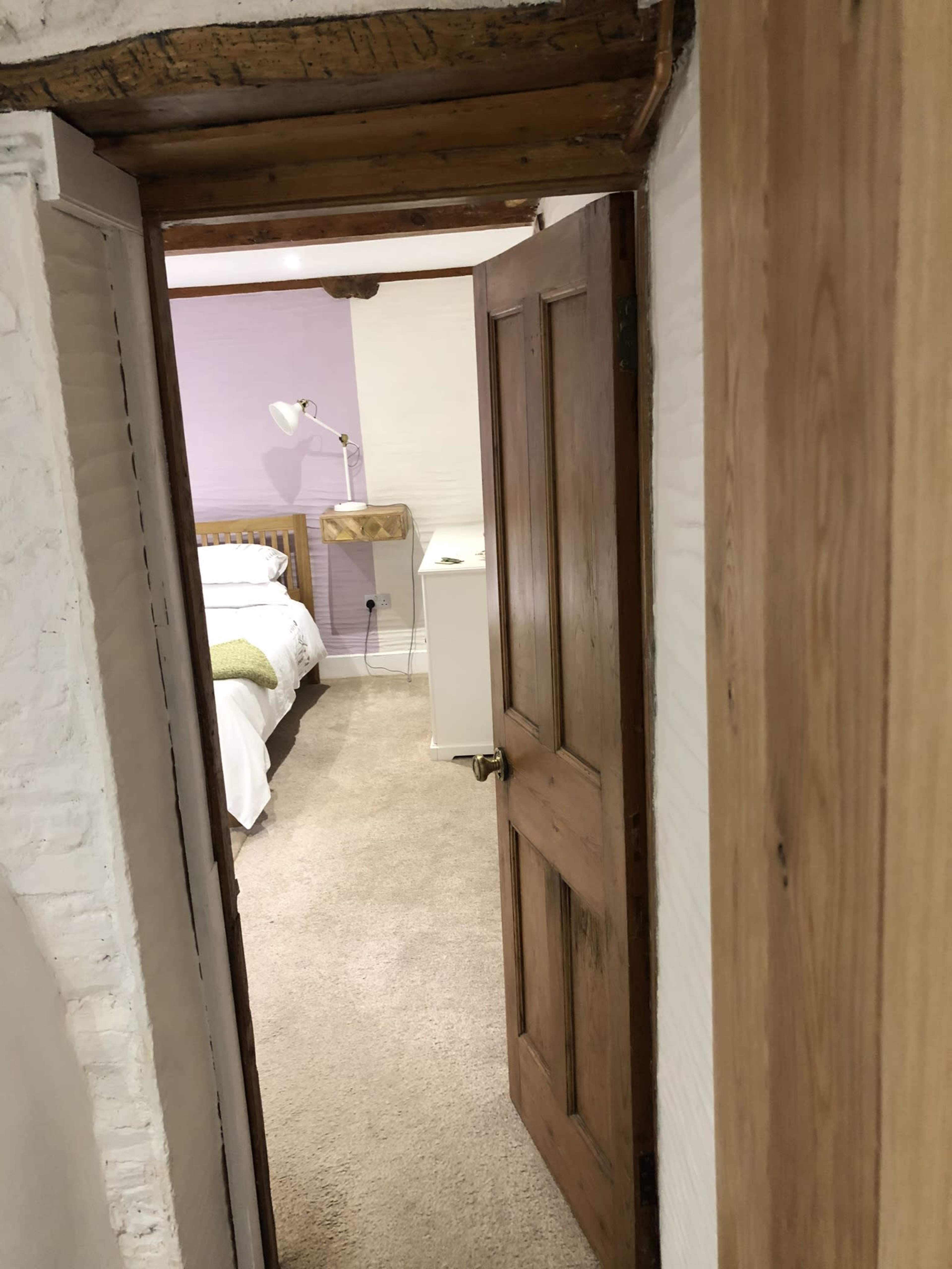 A wooden door opens into a softly lit bedroom featuring a bed, nightstand, and a lamp.