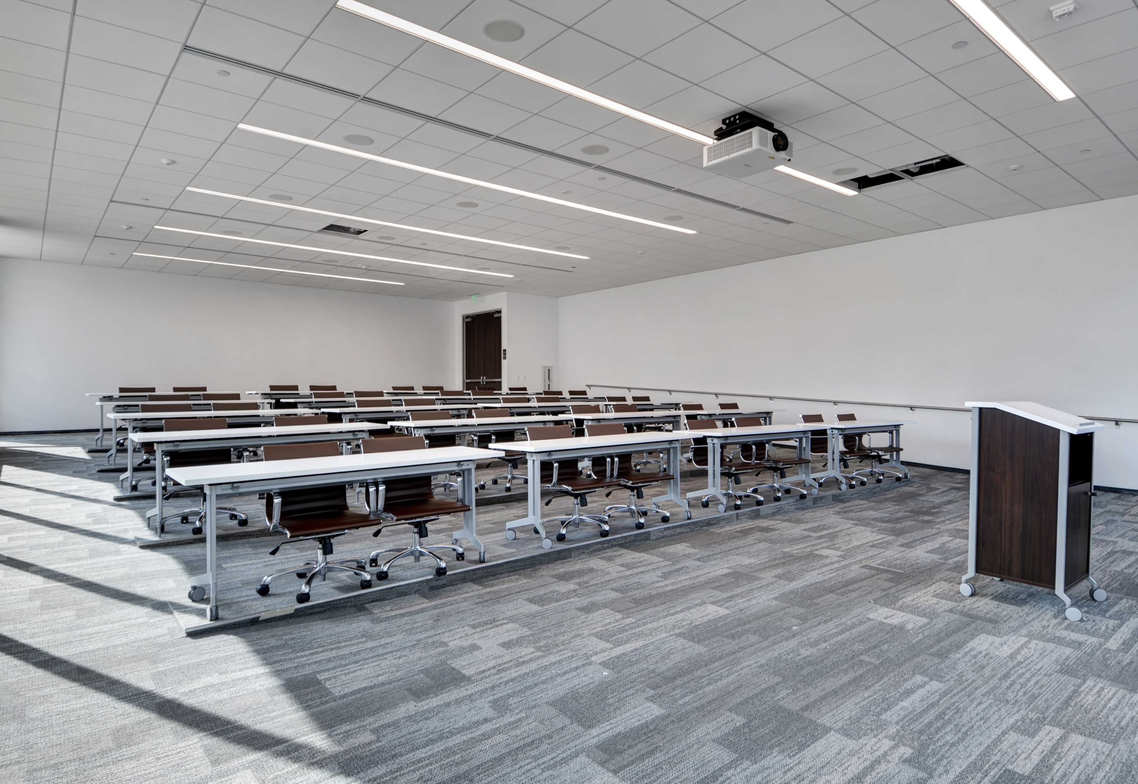 The image shows a modern classroom setup with rows of desks and chairs arranged for a presentation, featuring a podium and a projector overhead.