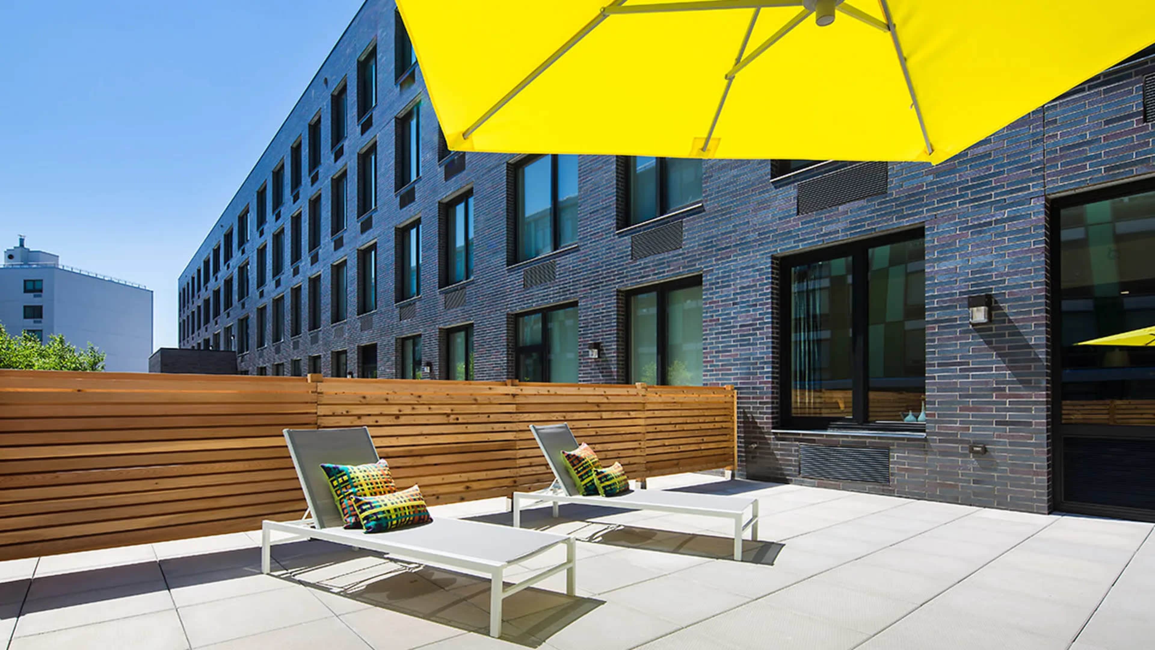 A terrace features two lounge chairs under a large yellow umbrella, surrounded by a wooden fence and brick building.