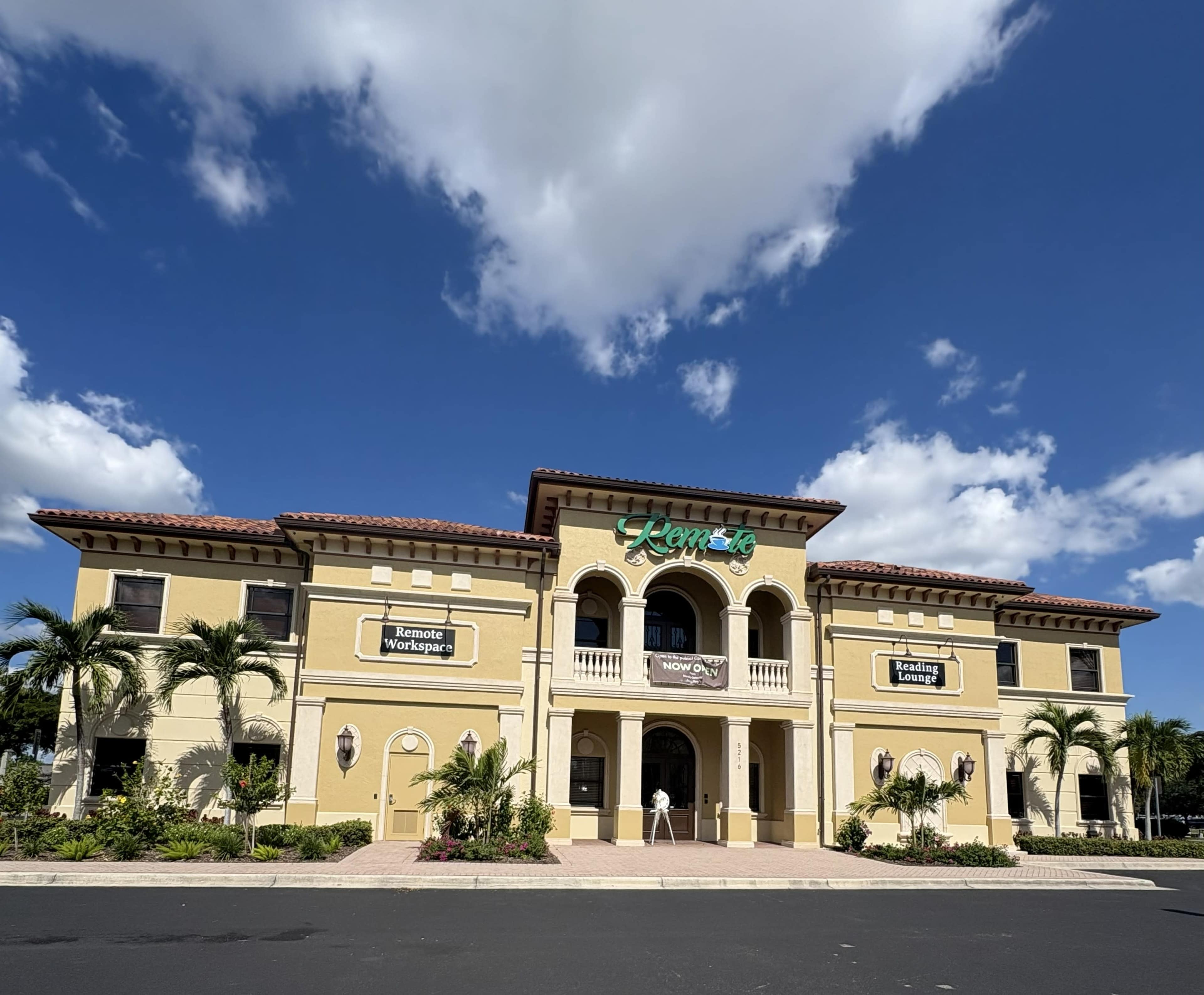 The image shows a large, two-story building with a distinctive architectural style, featuring palm trees in the foreground and a blue sky with clouds above.