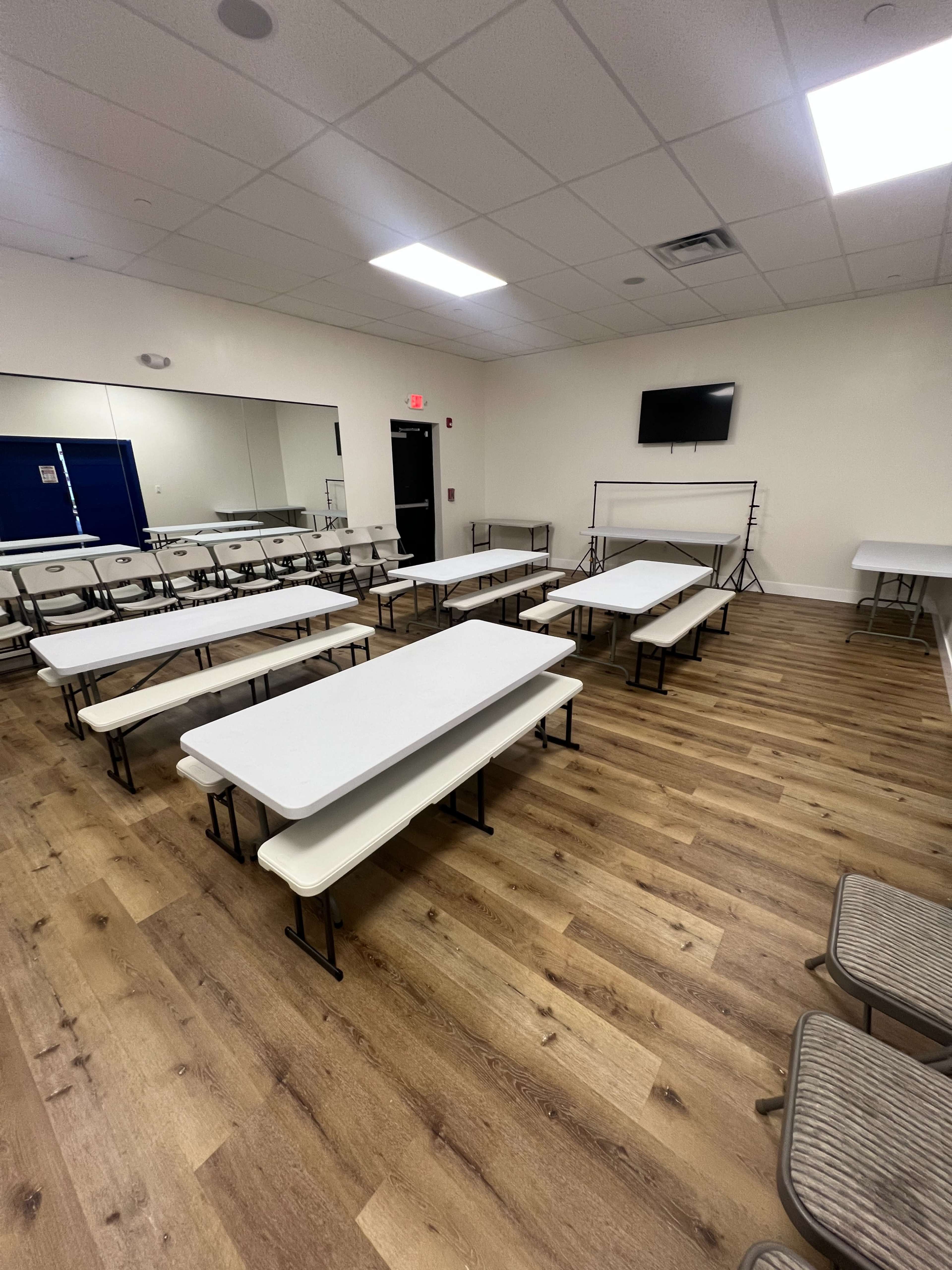 The image shows a spacious room with several white folding tables arranged in a grid pattern and wooden flooring, equipped with a mirror and a wall-mounted television.