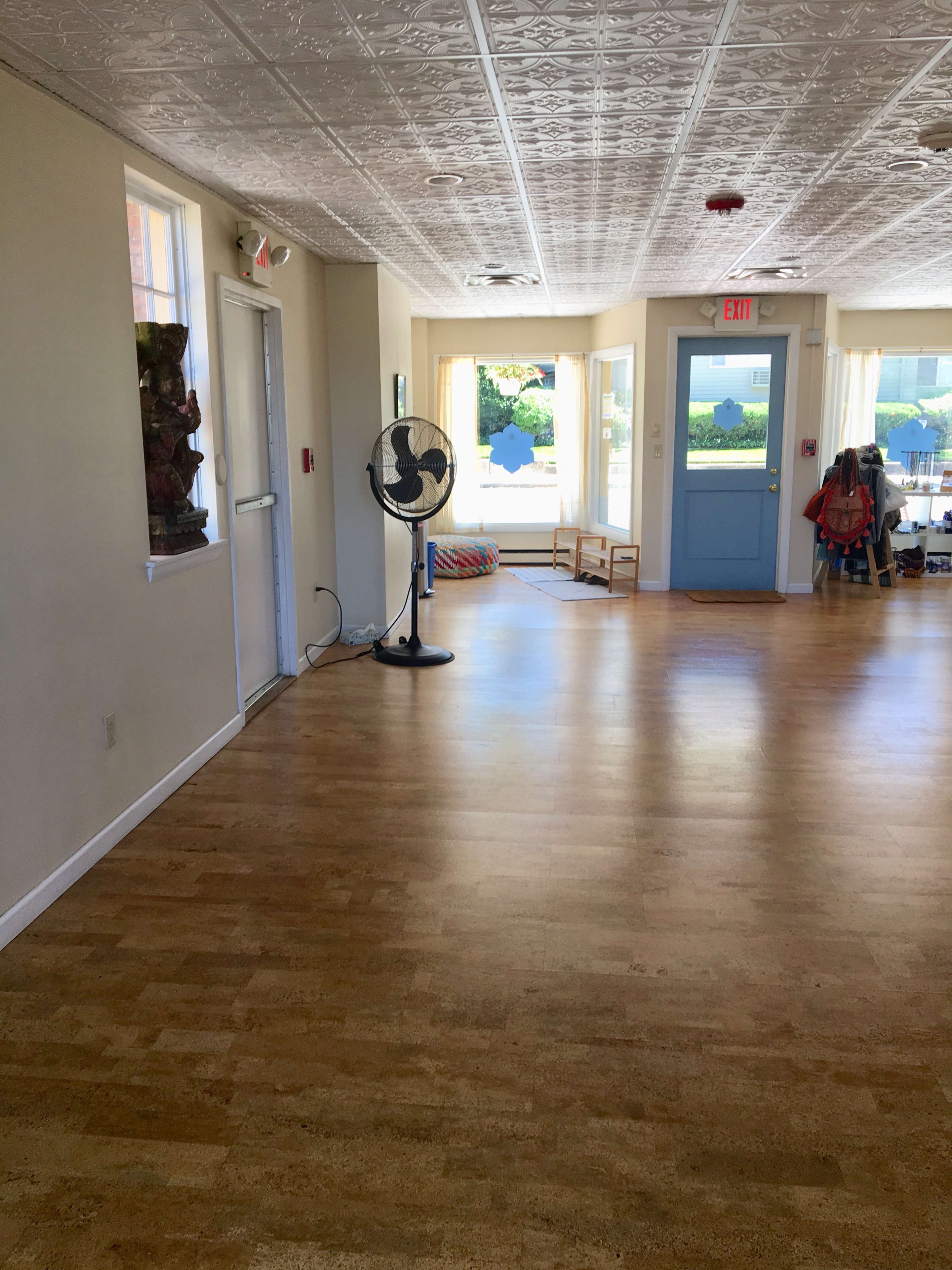 A spacious, brightly lit interior with wooden flooring, a fan, and double doors leading outside, flanked by large windows.