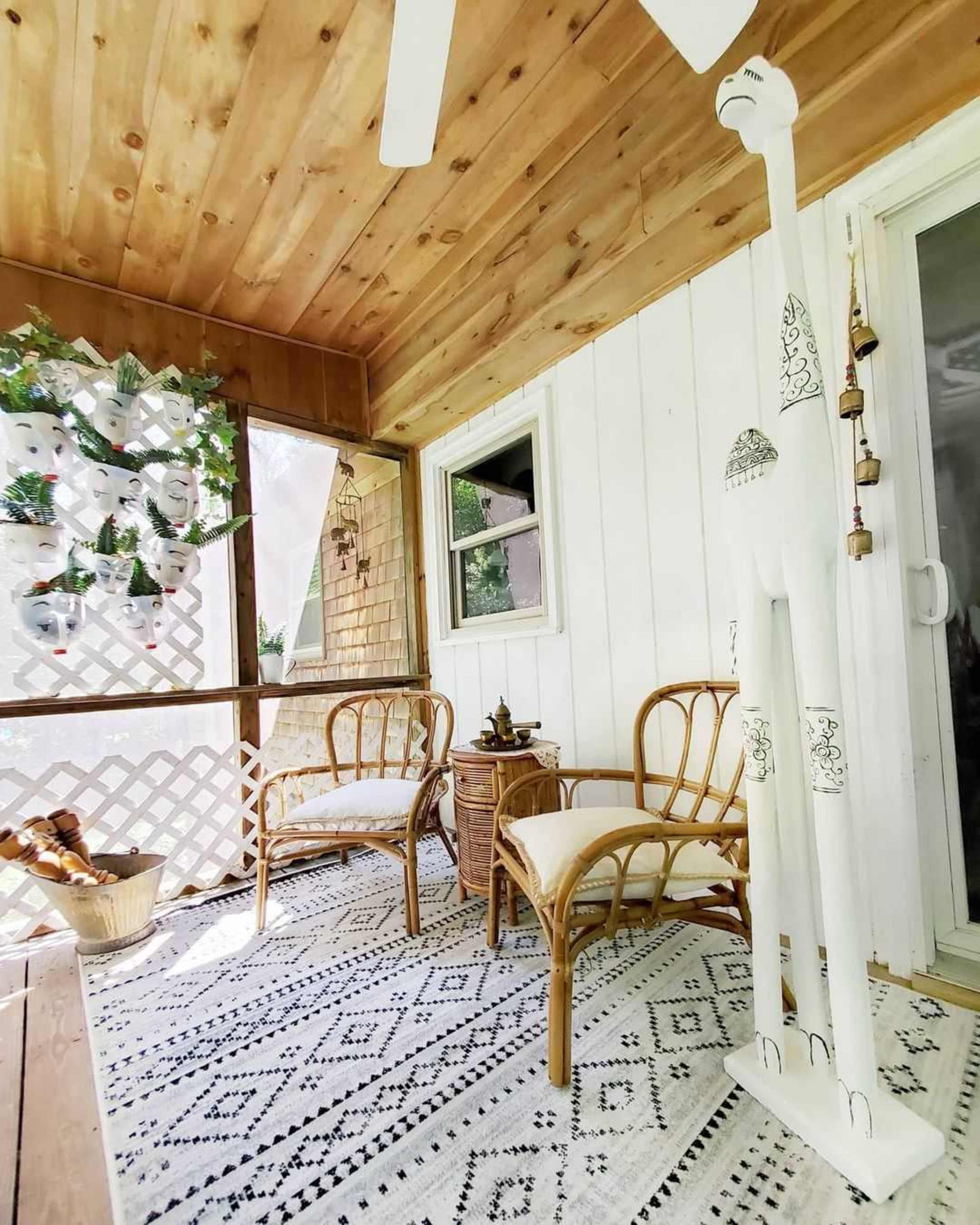 A sunlit porch featuring two rattan chairs, a small side table, and a tall, decorative giraffe, with a backdrop of a plant-laden wall and a wooden ceiling.