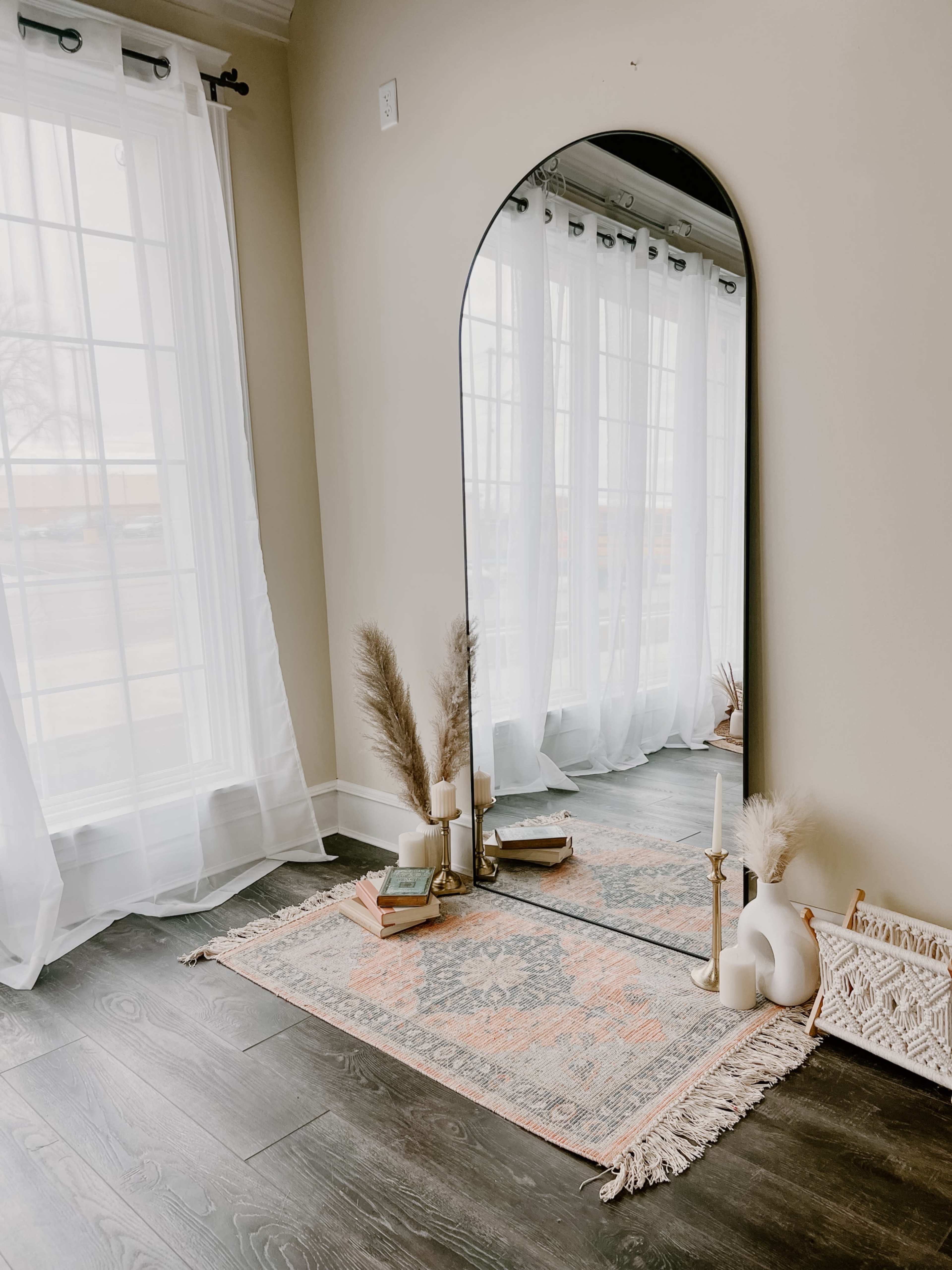 A large arched mirror stands against a wall near a window, reflecting a decorative rug and a few accessories on the floor.