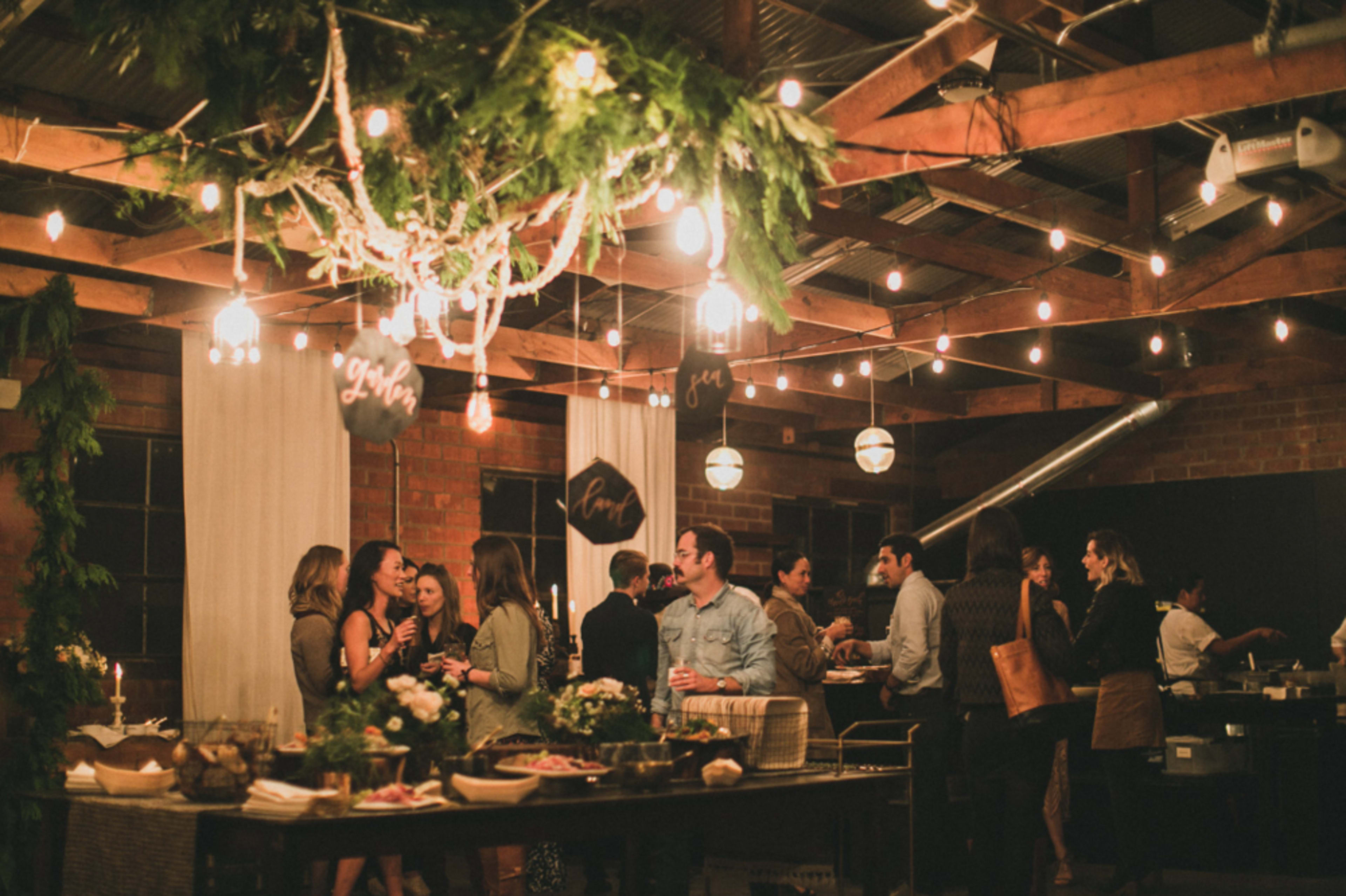 The image shows a festive gathering in a rustic space with string lights, people mingling, and a variety of food displayed on long tables.