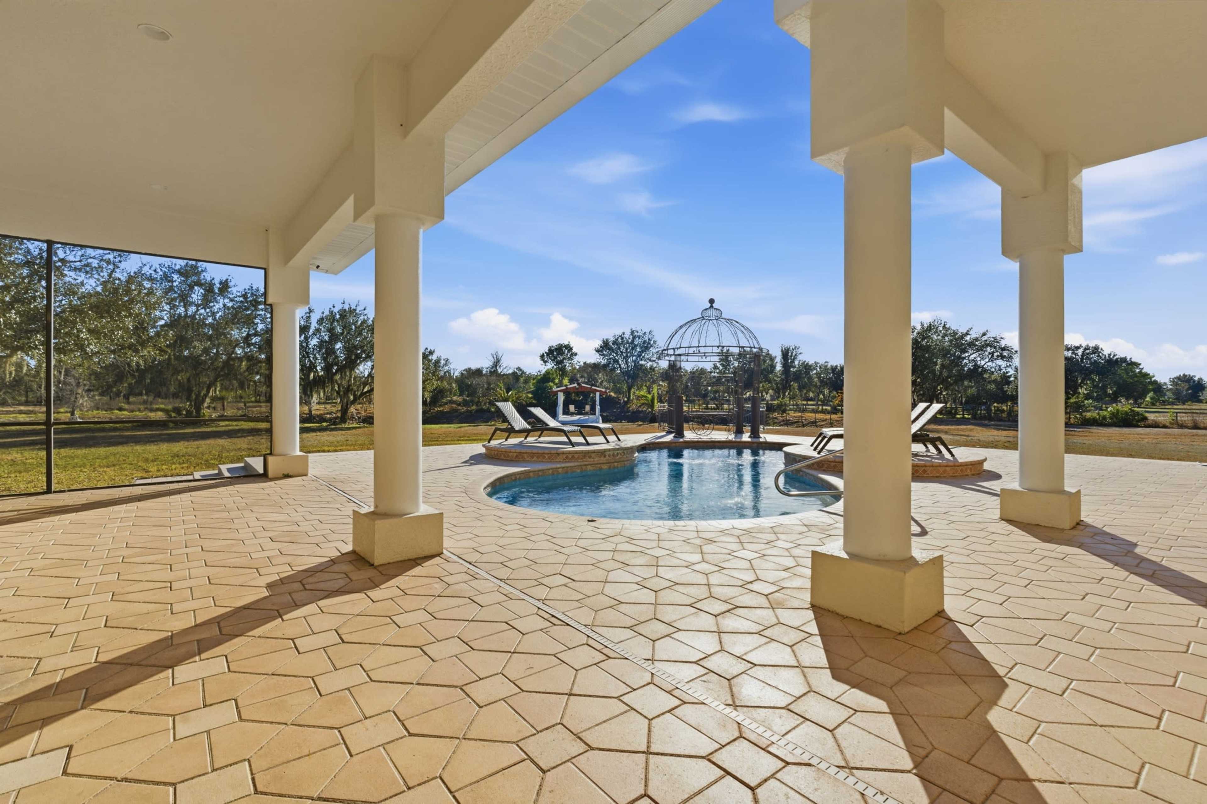The image shows a pool area with a circular pool, lounging chairs, and a decorative structure in a spacious outdoor setting surrounded by greenery.