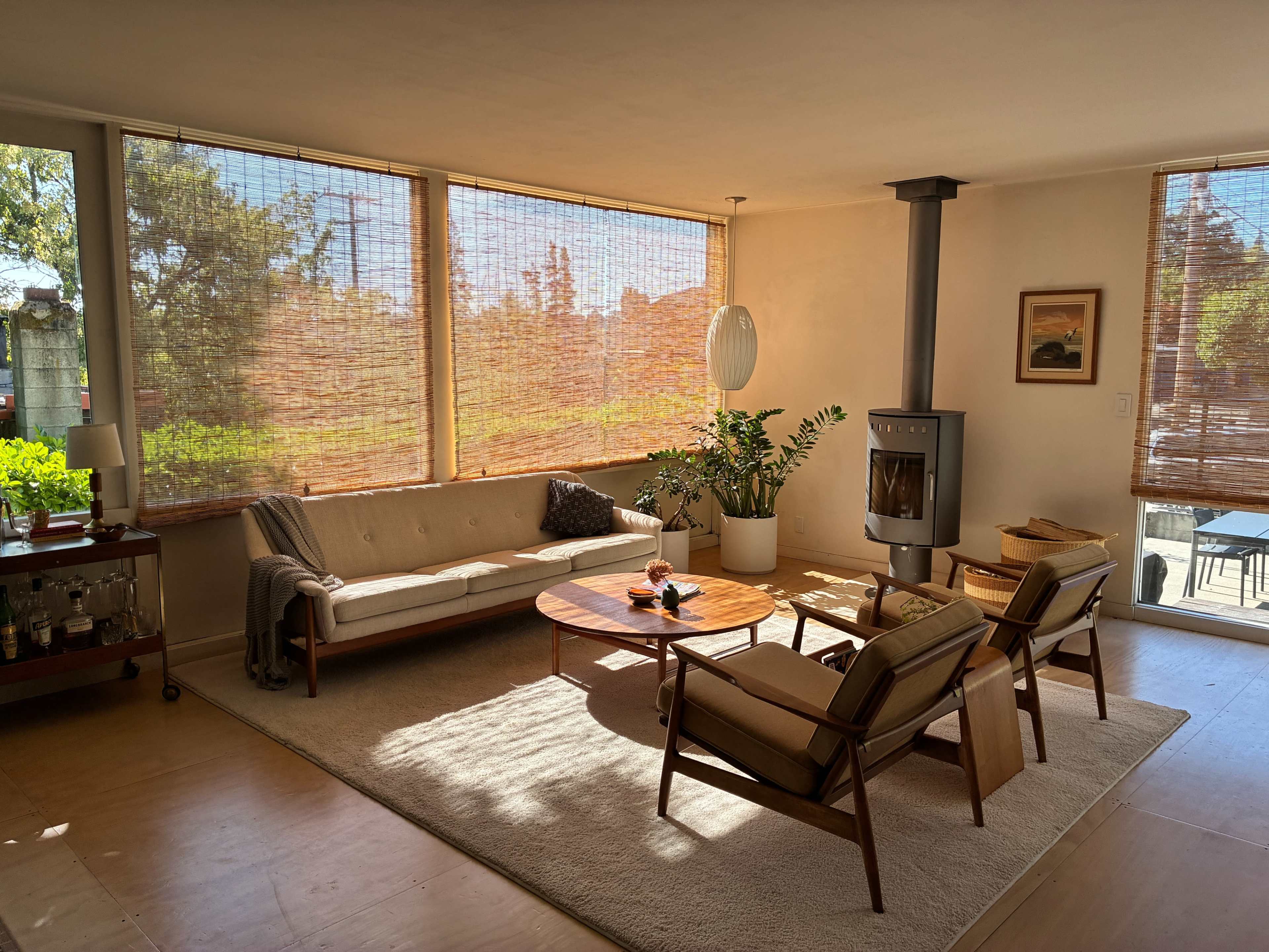 The image shows a modern living room featuring a light-colored sofa, two chairs, a circular coffee table, and a wood-burning stove, with large windows covered by woven shades.