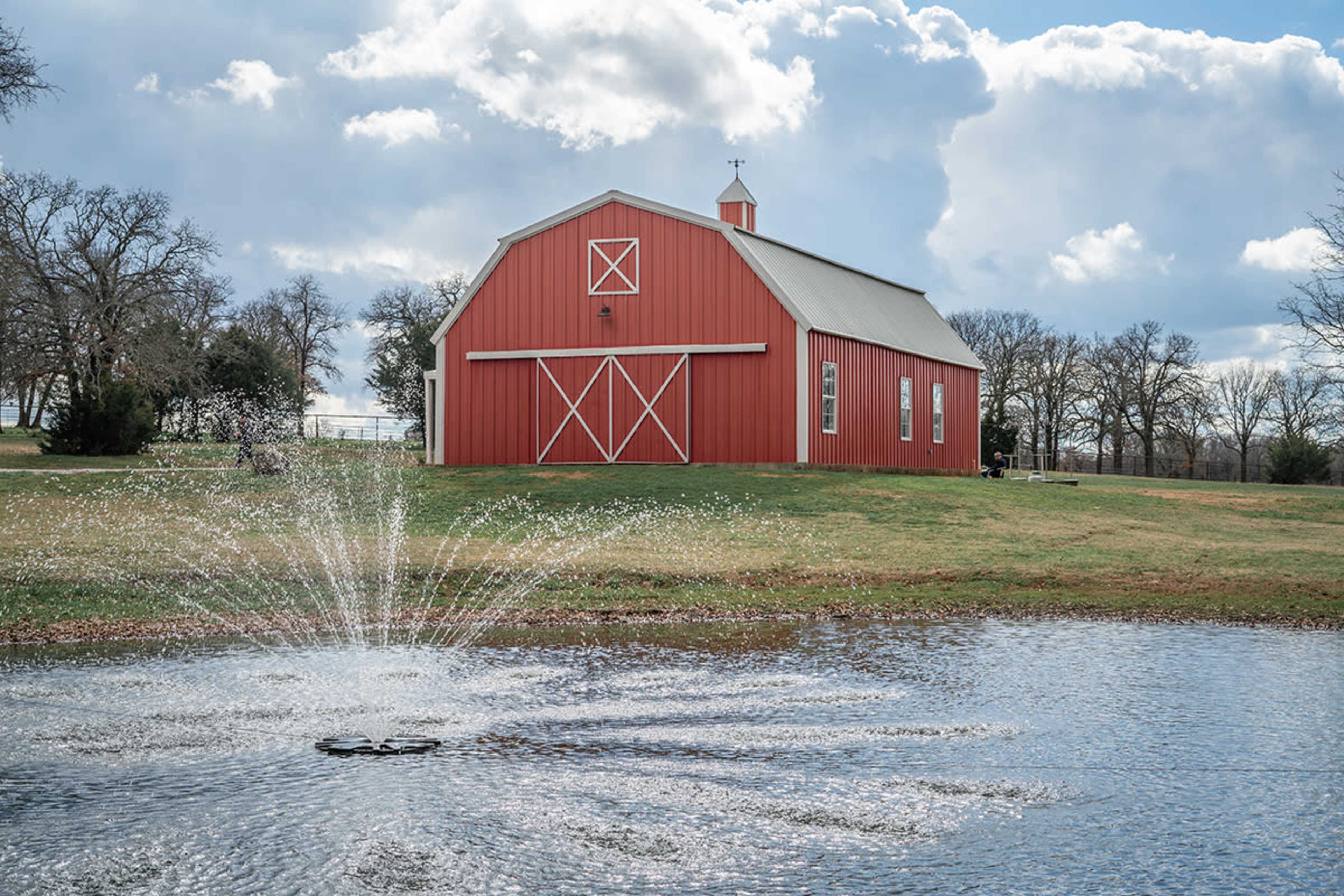 A red barn stands beside a pond with a fountain, surrounded by trees and a cloudy sky.
