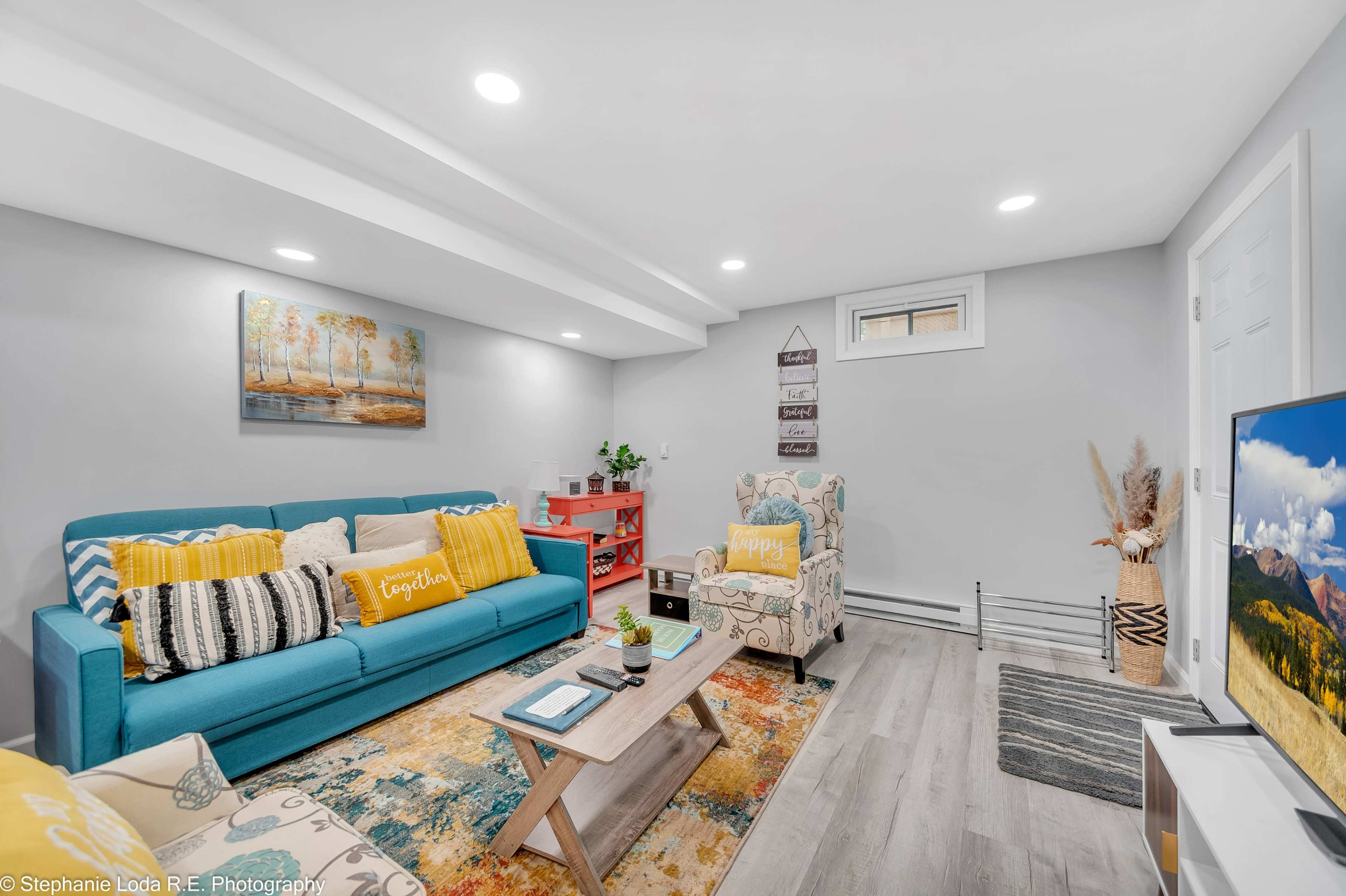 A cozy basement living area with a blue couch, decorative pillows, a wooden coffee table, and a patterned rug, complemented by light-colored walls and overhead lighting.
