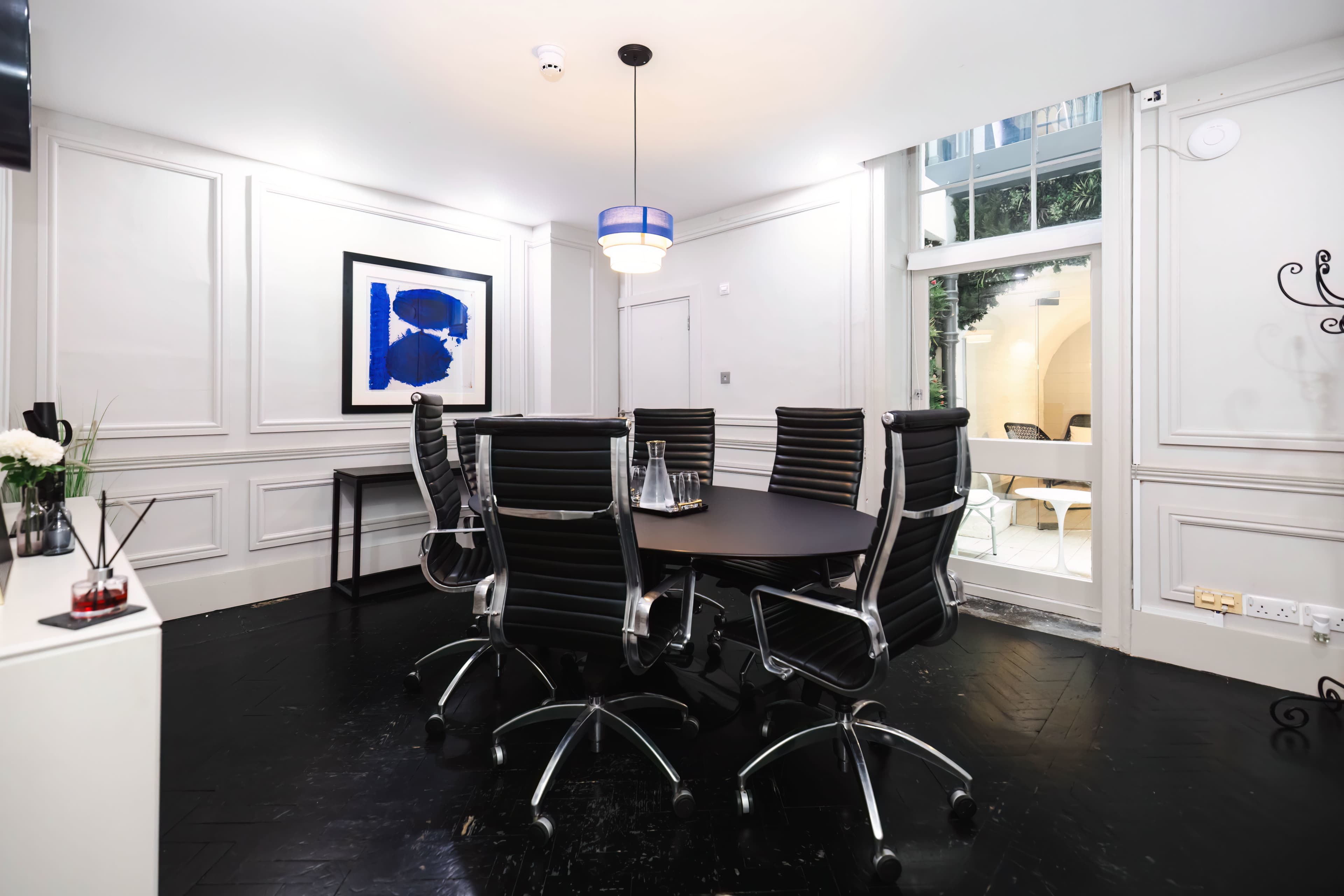 A modern conference room features a round table surrounded by black ergonomic chairs, with large windows allowing natural light to enter, and a piece of abstract art on the wall.