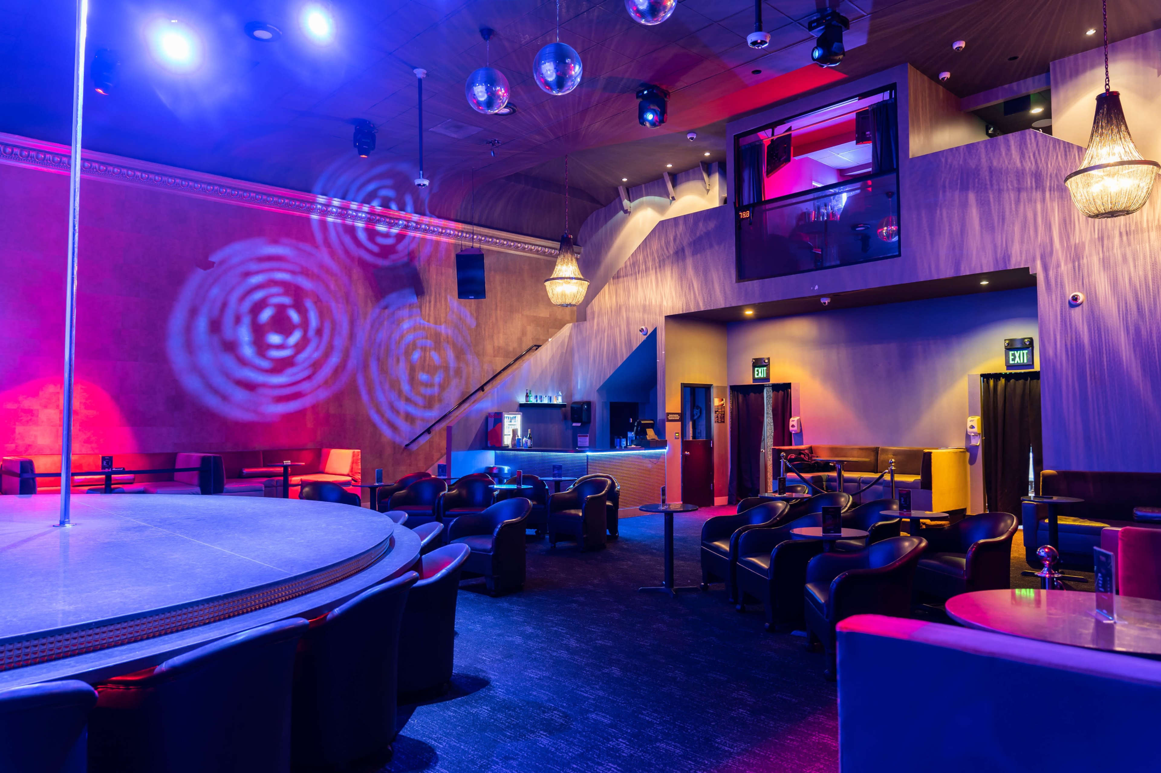 The interior of a dimly lit club features a dance pole at the center, surrounded by circular seating and colorful lighting effects on the walls.