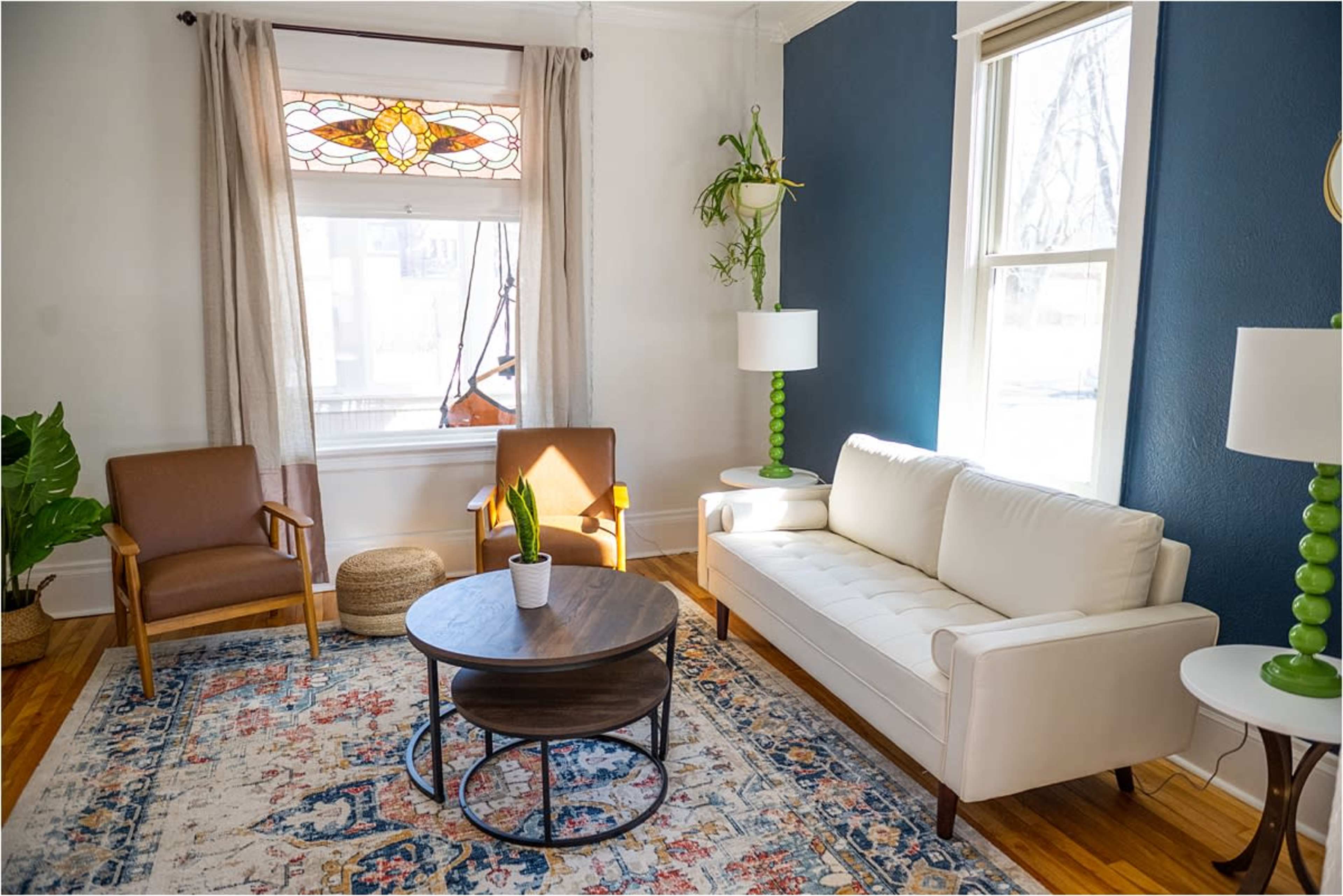 The living room features a white sofa, two brown armchairs, a round coffee table, and large windows with sheer curtains.
