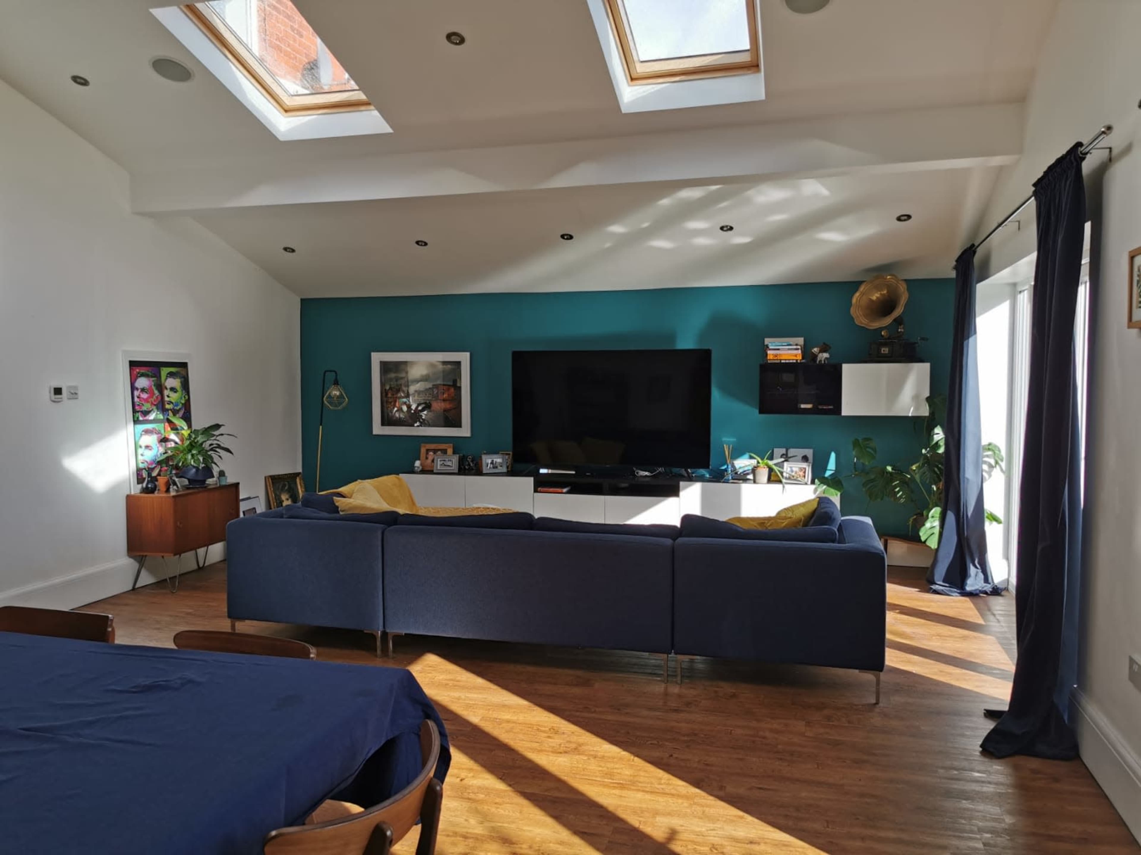 A modern living room with a blue sectional sofa, a large television on a teal wall, skylights providing natural light, and a dining table in the foreground.