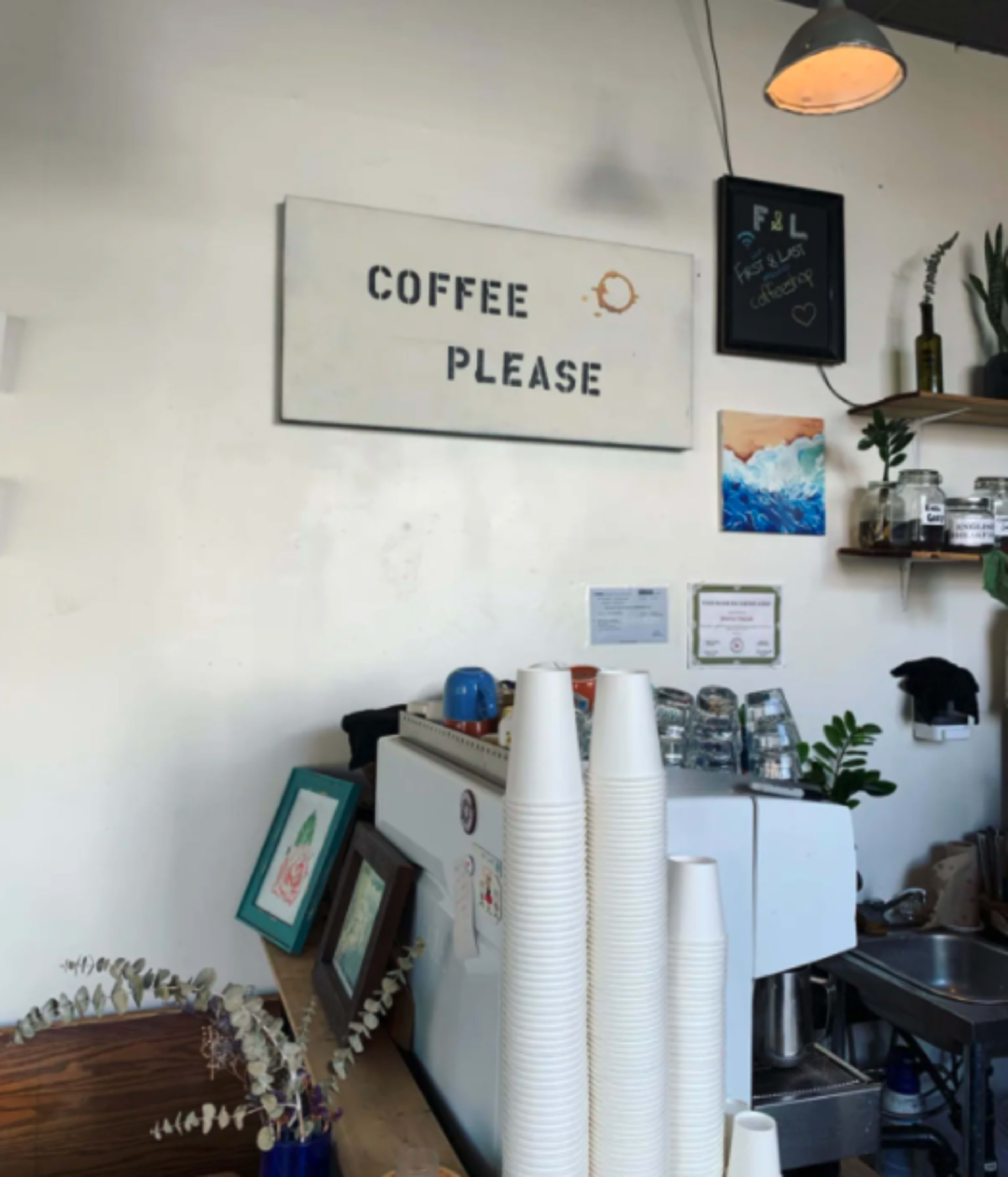 A coffee shop interior features a sign that reads "COFFEE PLEASE" above a white coffee machine, several cups stacked neatly, and decorative plants.