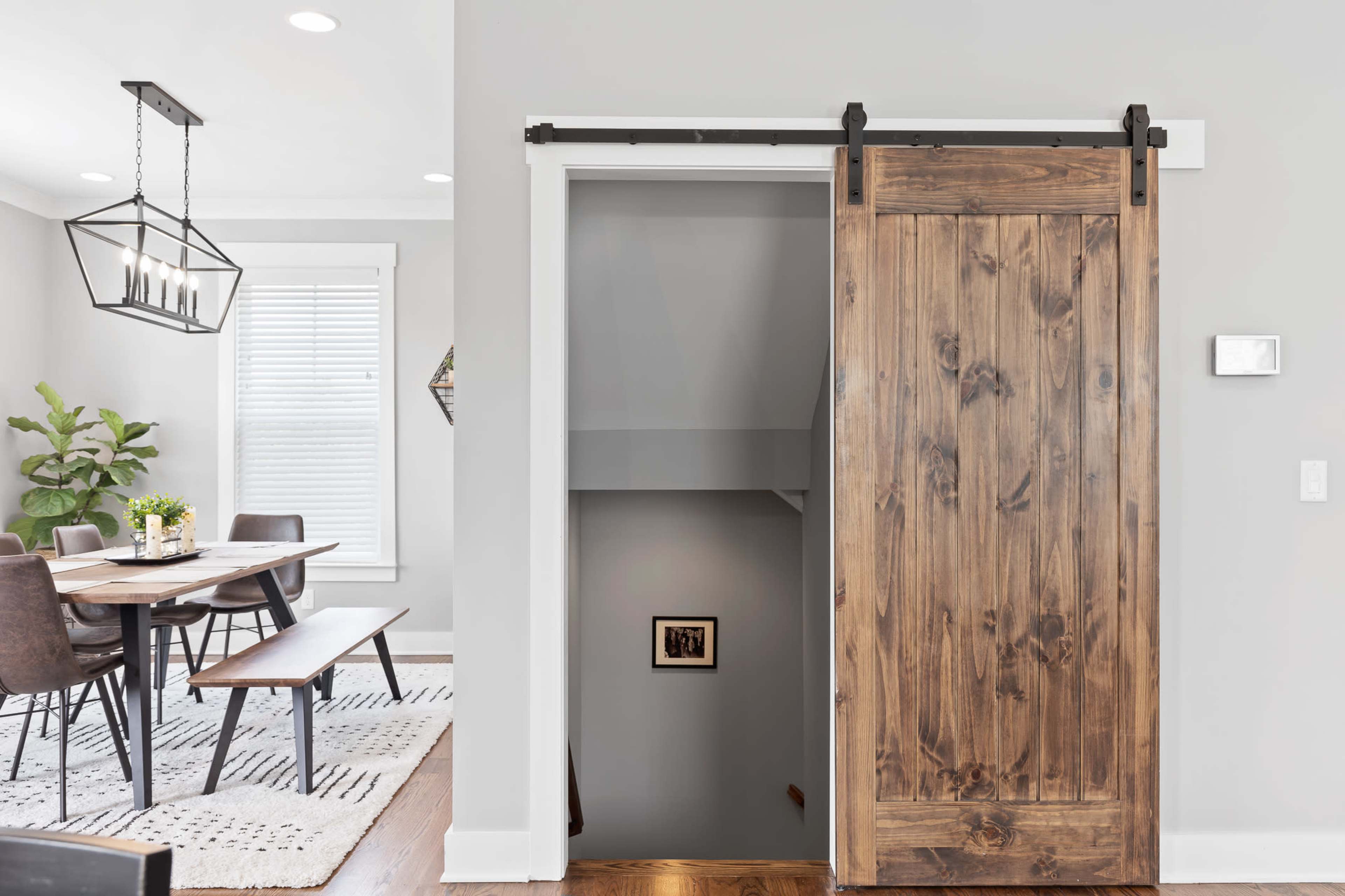 A sliding wooden barn door partially opens to reveal a staircase leading down, adjacent to a dining area with a long table and chairs.