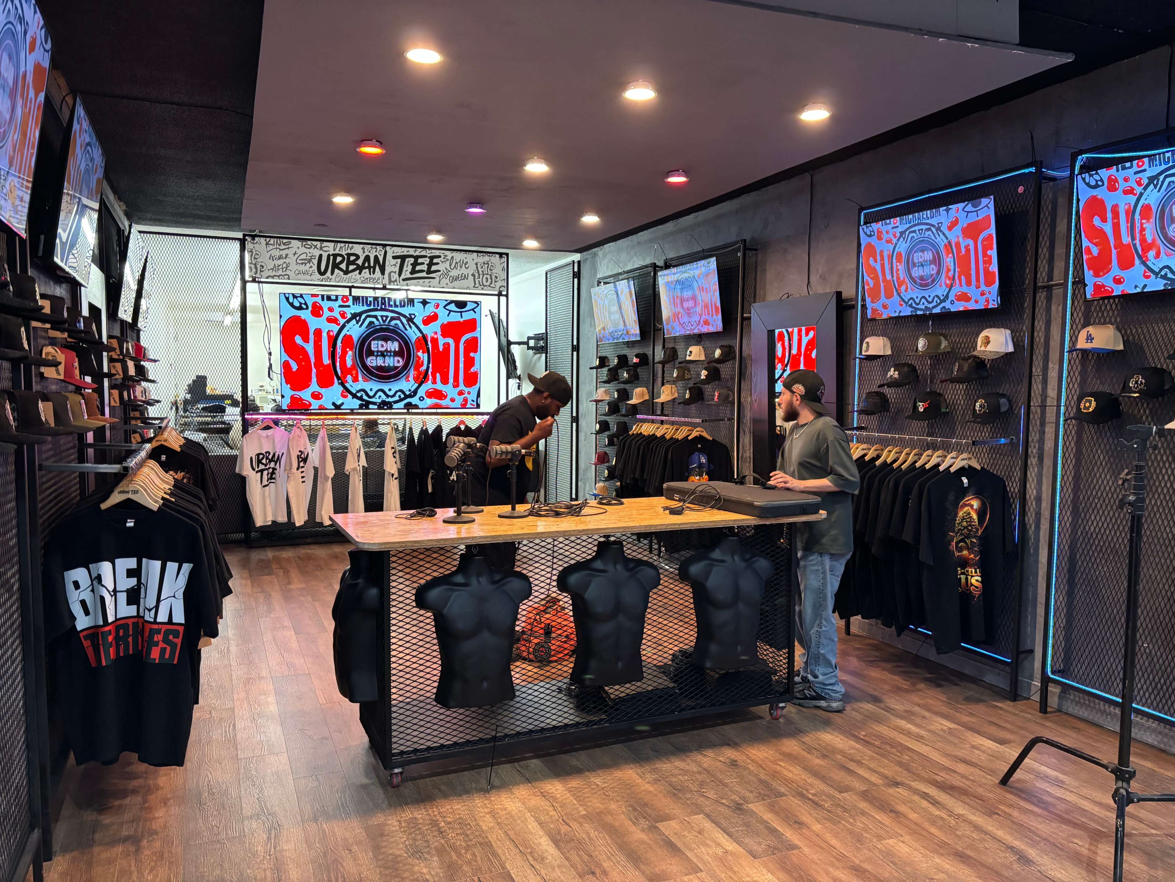 The image shows a clothing retail store with racks displaying various t-shirts and hats, and a central checkout counter where two individuals are engaged in conversation.