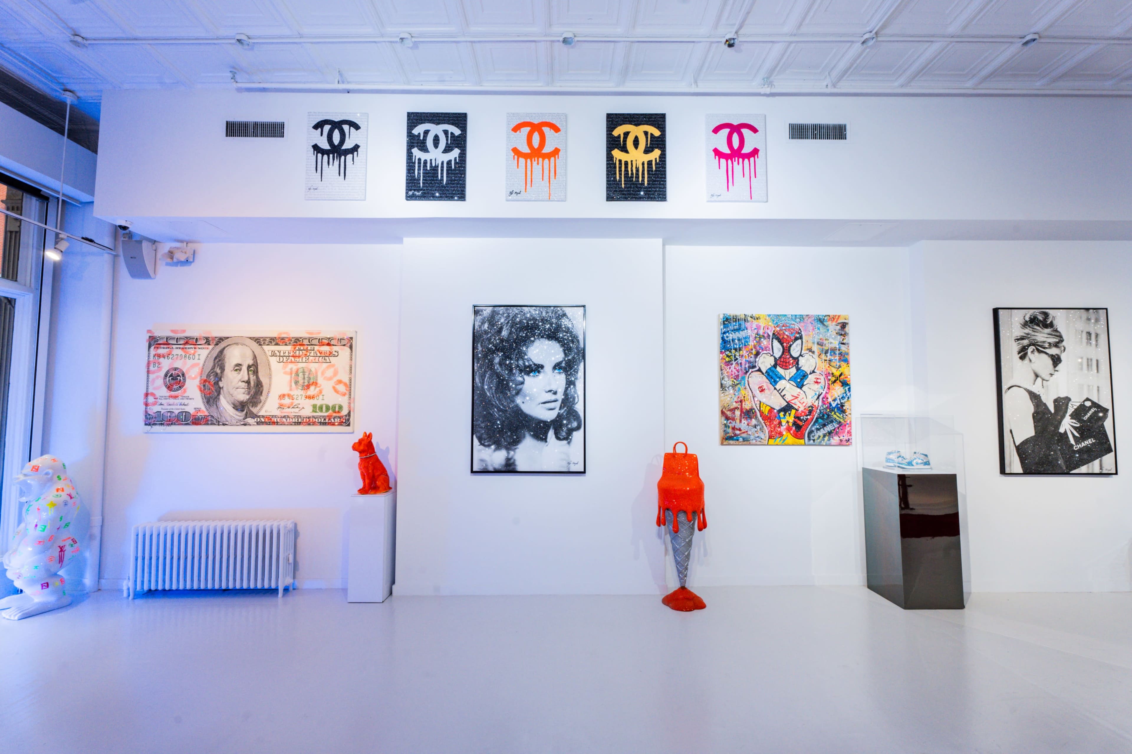 The image shows an art gallery with various framed artworks on white walls, featuring a mix of prints and paintings, along with sculptures displayed on the floor.