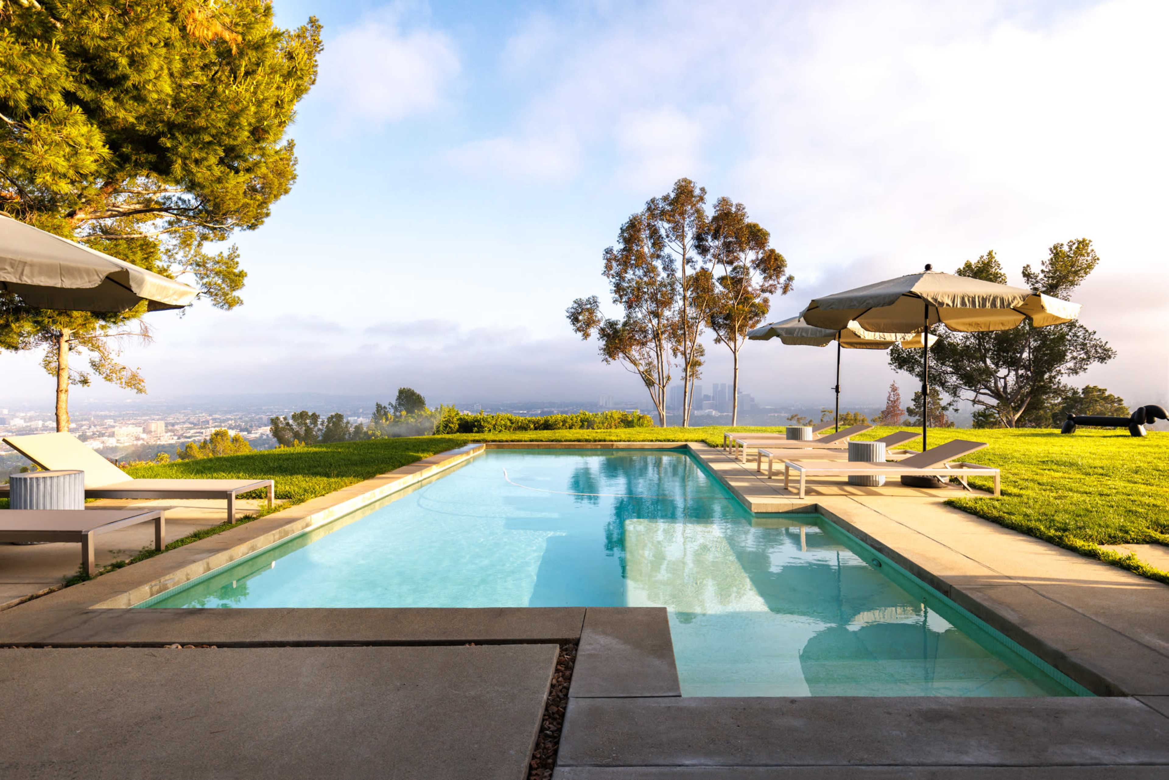 A modern swimming pool is situated on a hillside with lounge chairs and umbrellas, overlooking a cityscape under a partly cloudy sky.