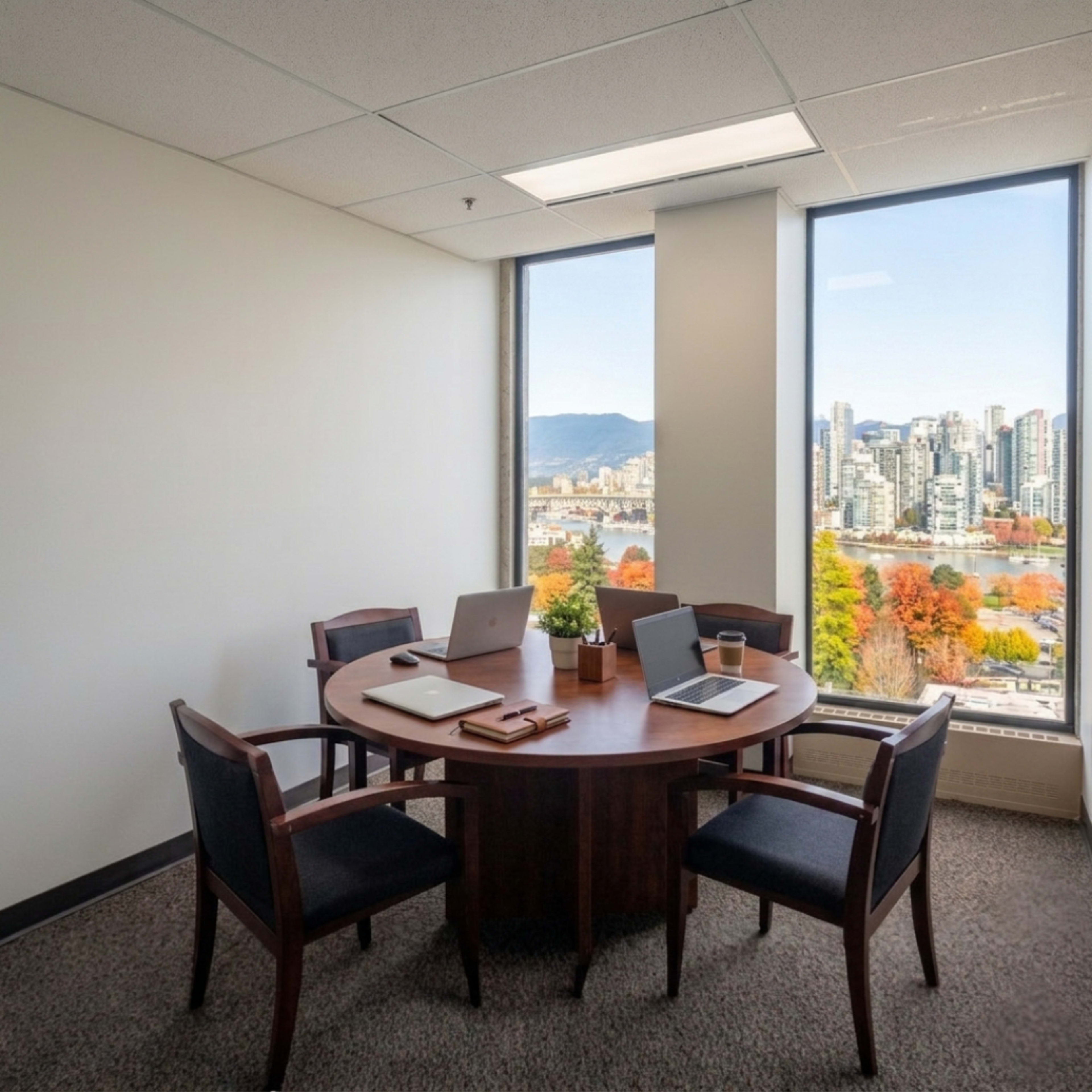 A modern office with a round wooden table and four chairs overlooks a cityscape and mountains through large windows.