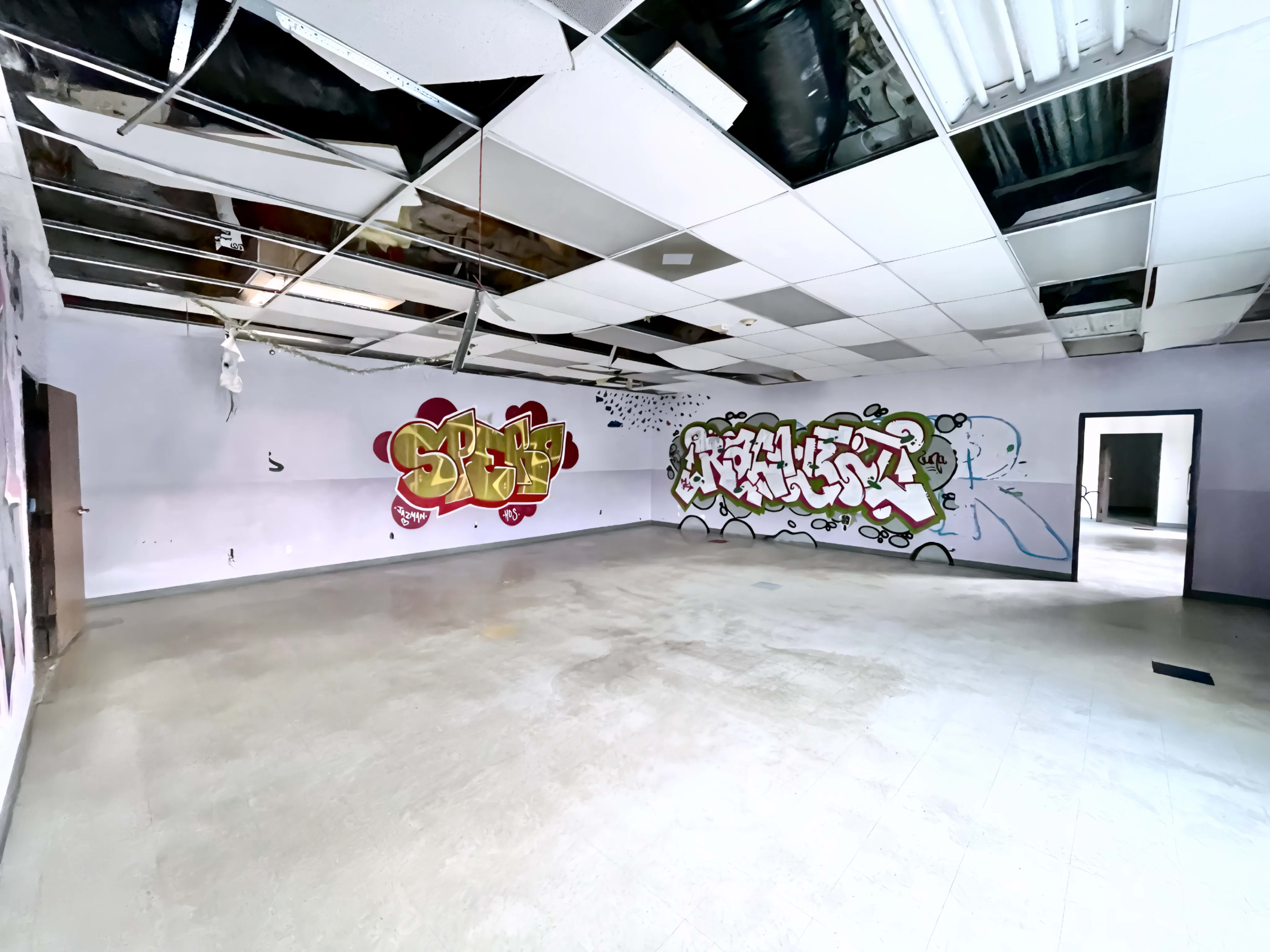 The image shows an empty room with graffiti on two walls and exposed ceiling tiles.