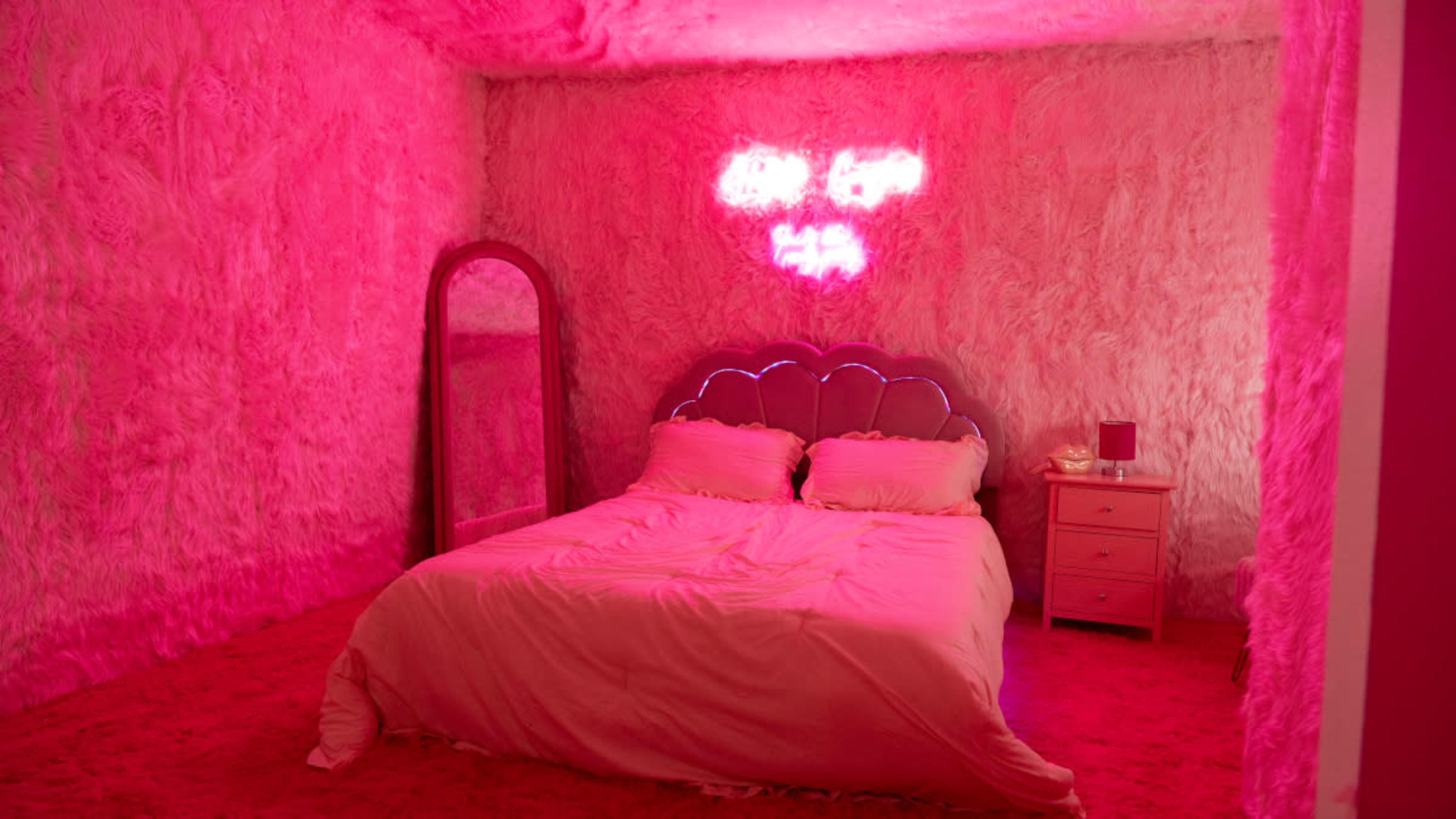 A pink-themed bedroom features a plush wall texture, a bed with a light-colored comforter, a mirror, and a bedside table.