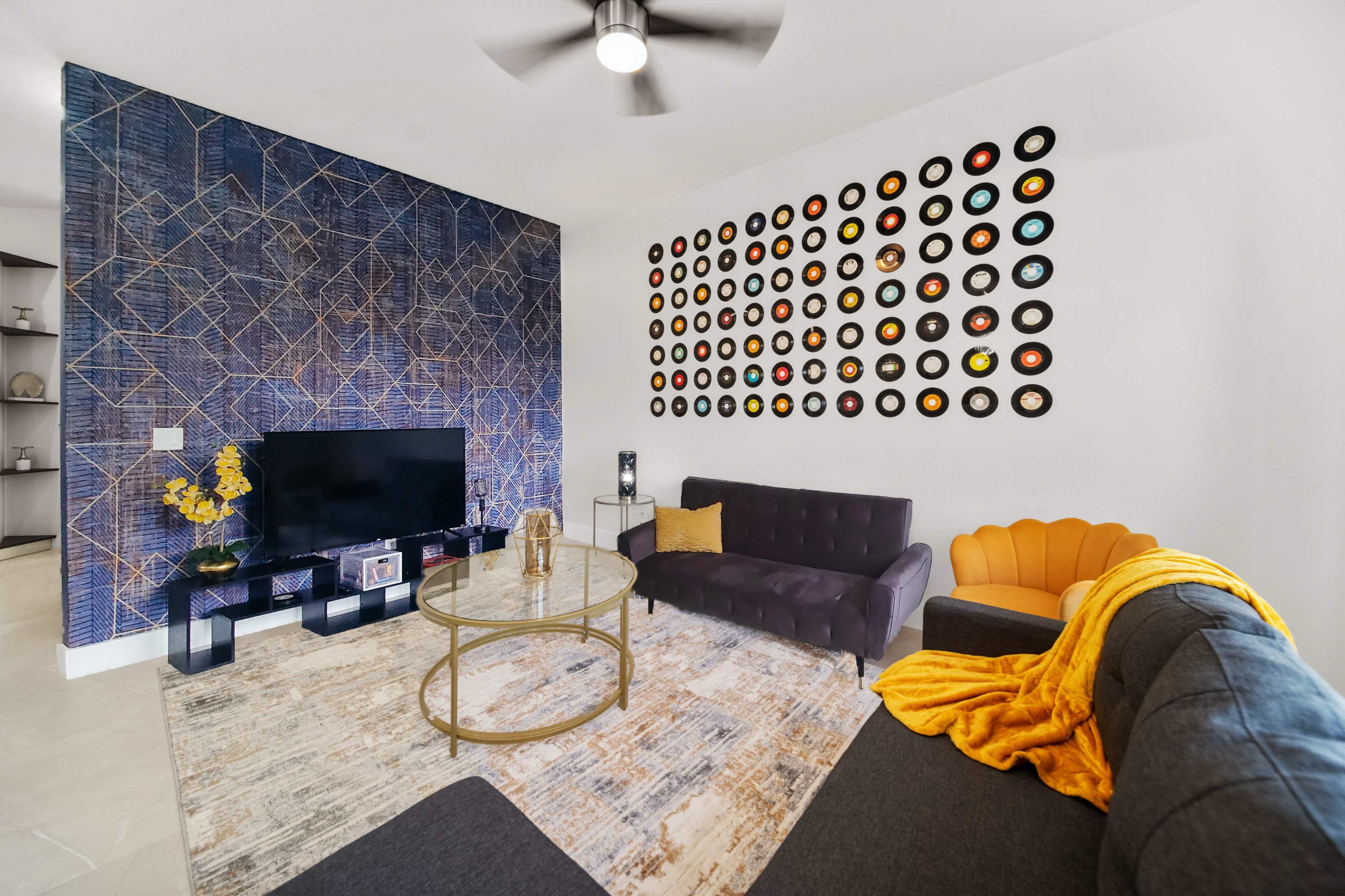 A modern living room featuring a television on a stand, a circular coffee table, a dark sofa, and an orange accent chair against a patterned wall adorned with vinyl records.