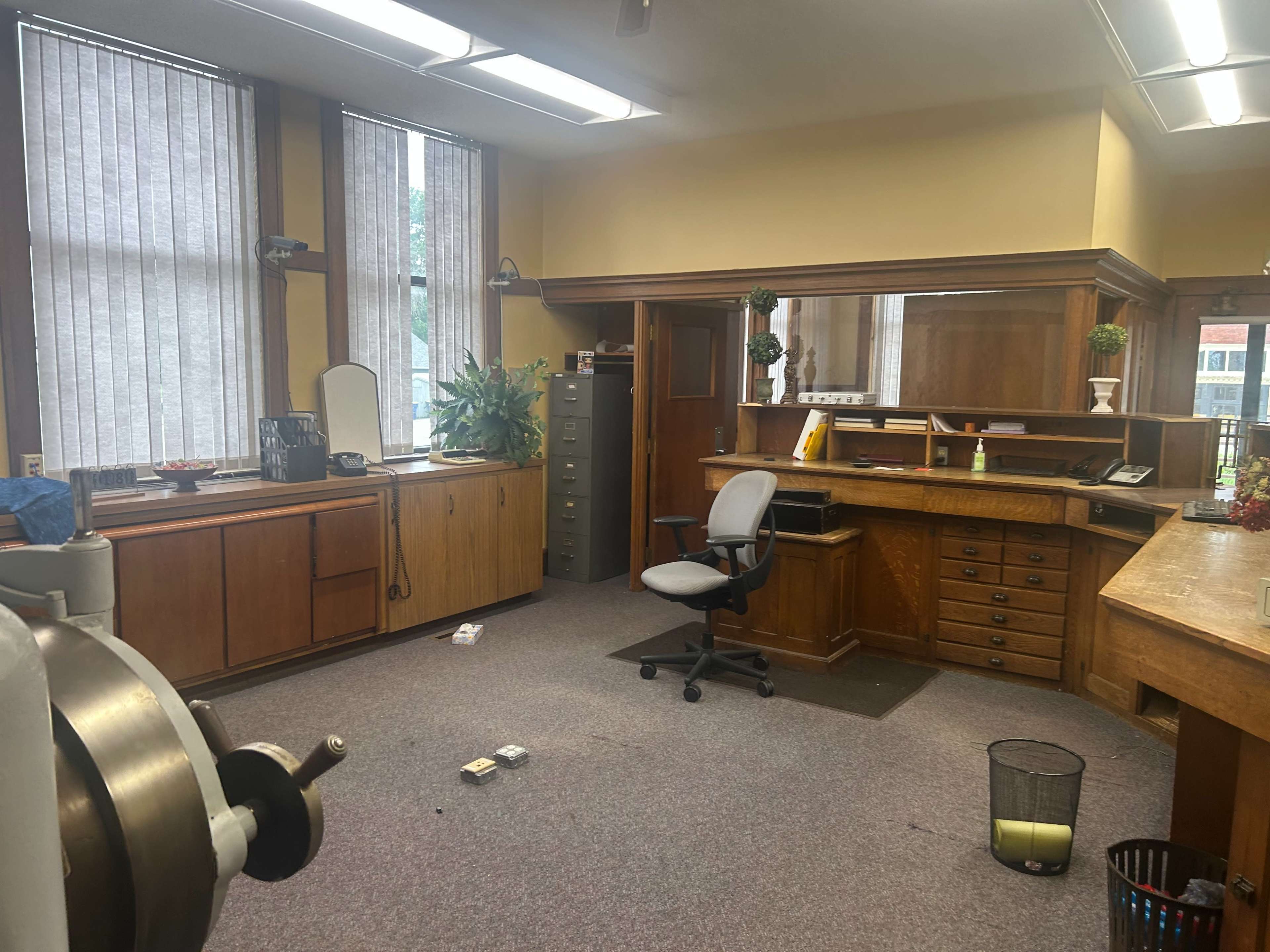 The image shows a spacious office with wooden furniture, including a desk and filing cabinet, along with a swivel chair and plants near the windows.
