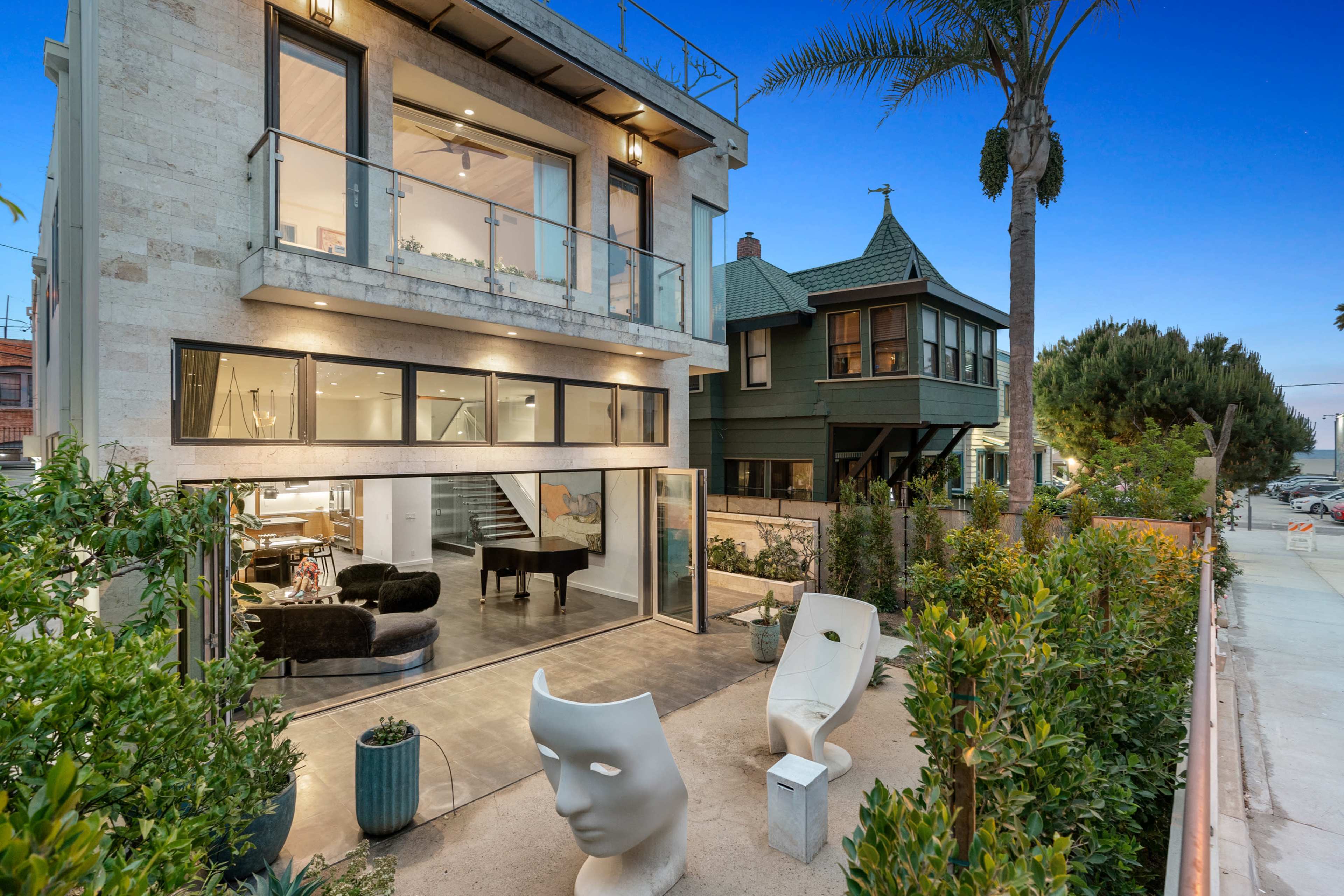 A modern two-story house with large windows and a balcony, surrounded by landscaped greenery and featuring a unique sculpture in the foreground.