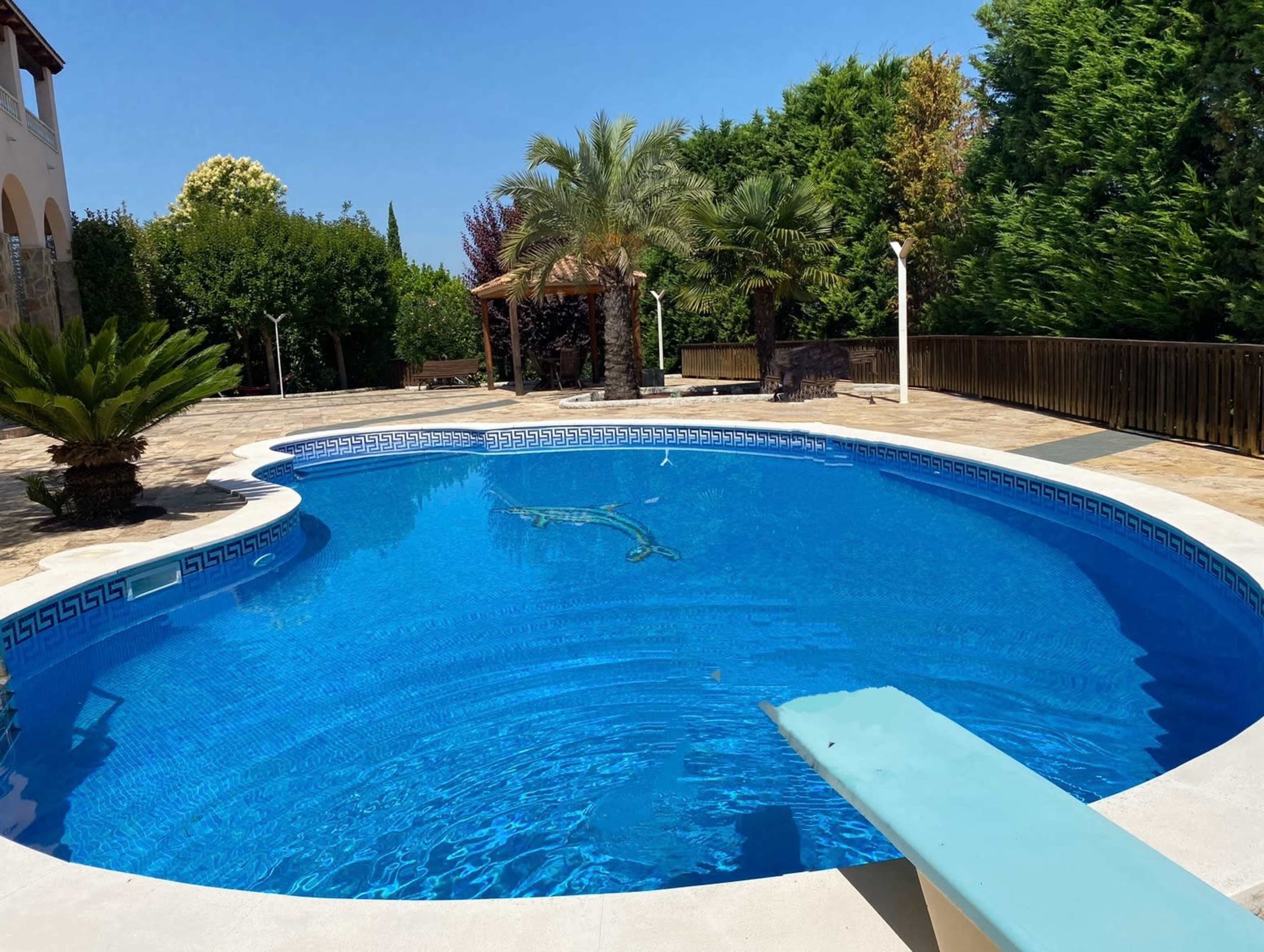 The image shows a blue swimming pool with a diving board surrounded by landscaped greenery and palm trees.