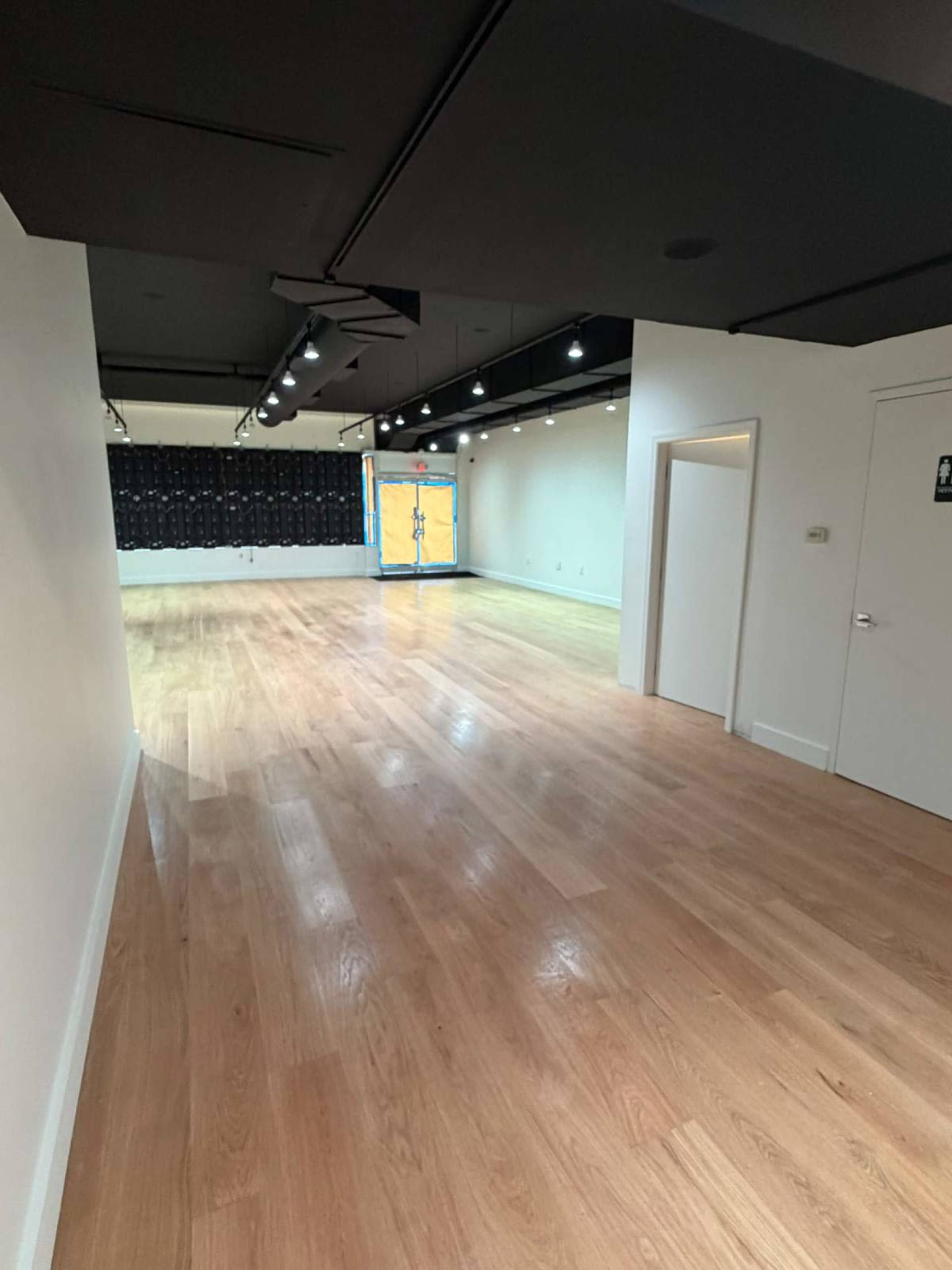 The image shows a spacious, empty room with light-colored wooden flooring, a black ceiling, and a wall featuring lockers at the far end.
