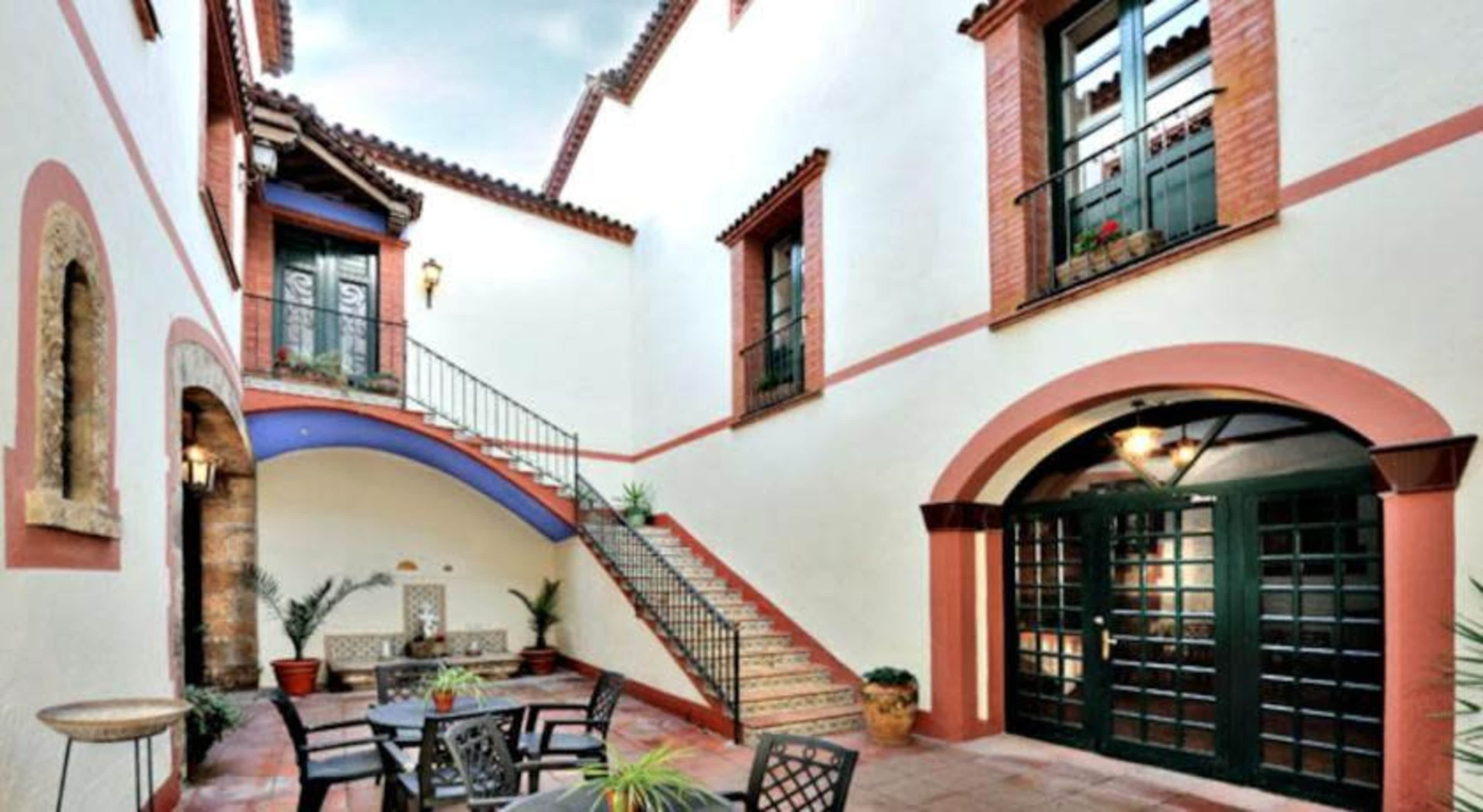 The image shows a courtyard with a staircase leading to upper levels, surrounded by white walls and red accents, while potted plants and outdoor furniture are positioned on the tiled floor.