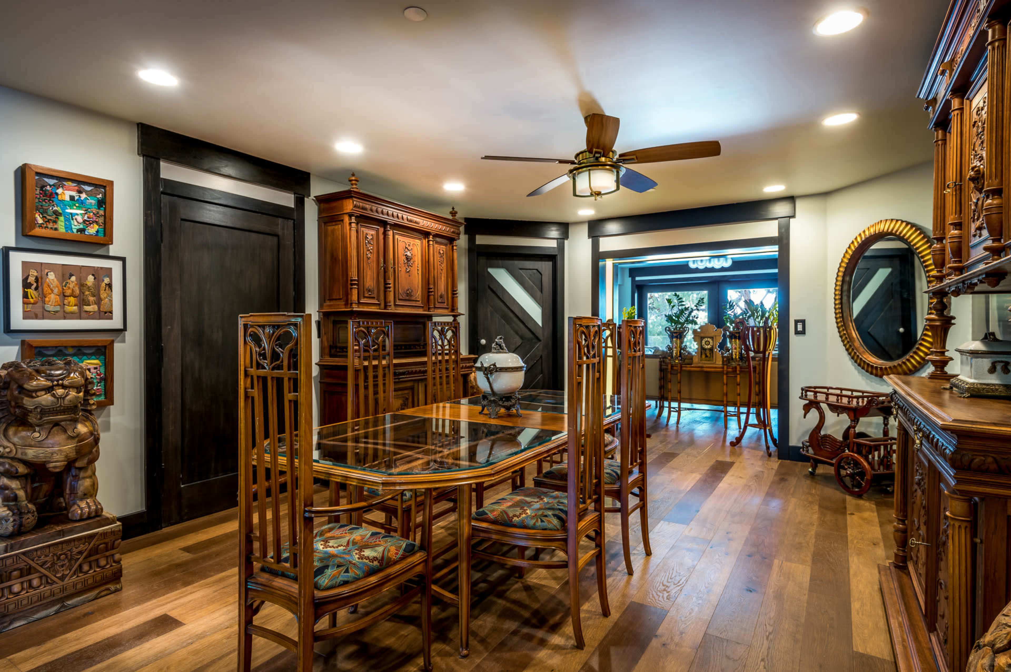 A dining room features a wooden table with chairs, decorative furniture, and a ceiling fan, leading to a brighter area with visible plants.