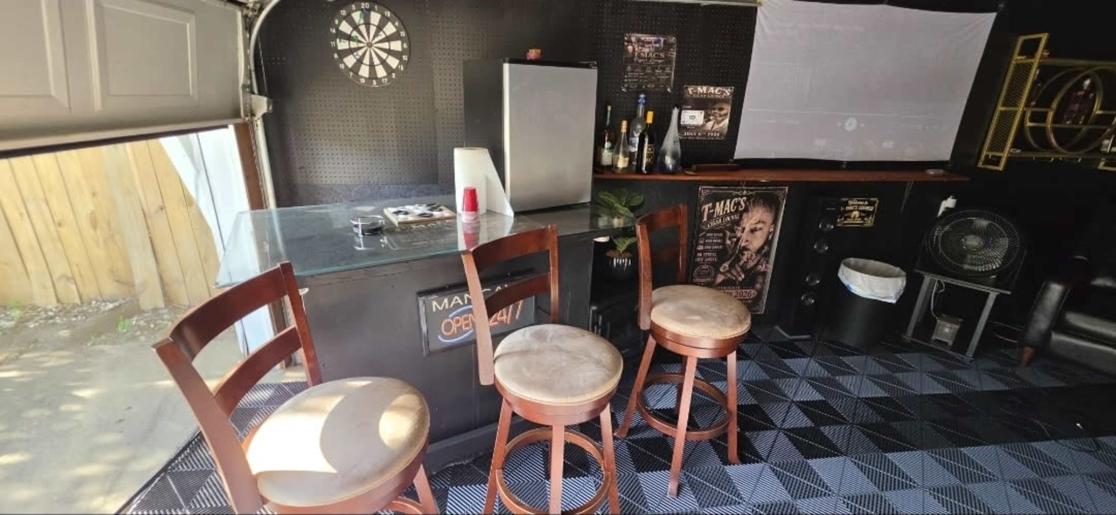 The image shows a garage bar area featuring a small countertop with three bar stools, a dartboard on the wall, and various bottles displayed on shelves.