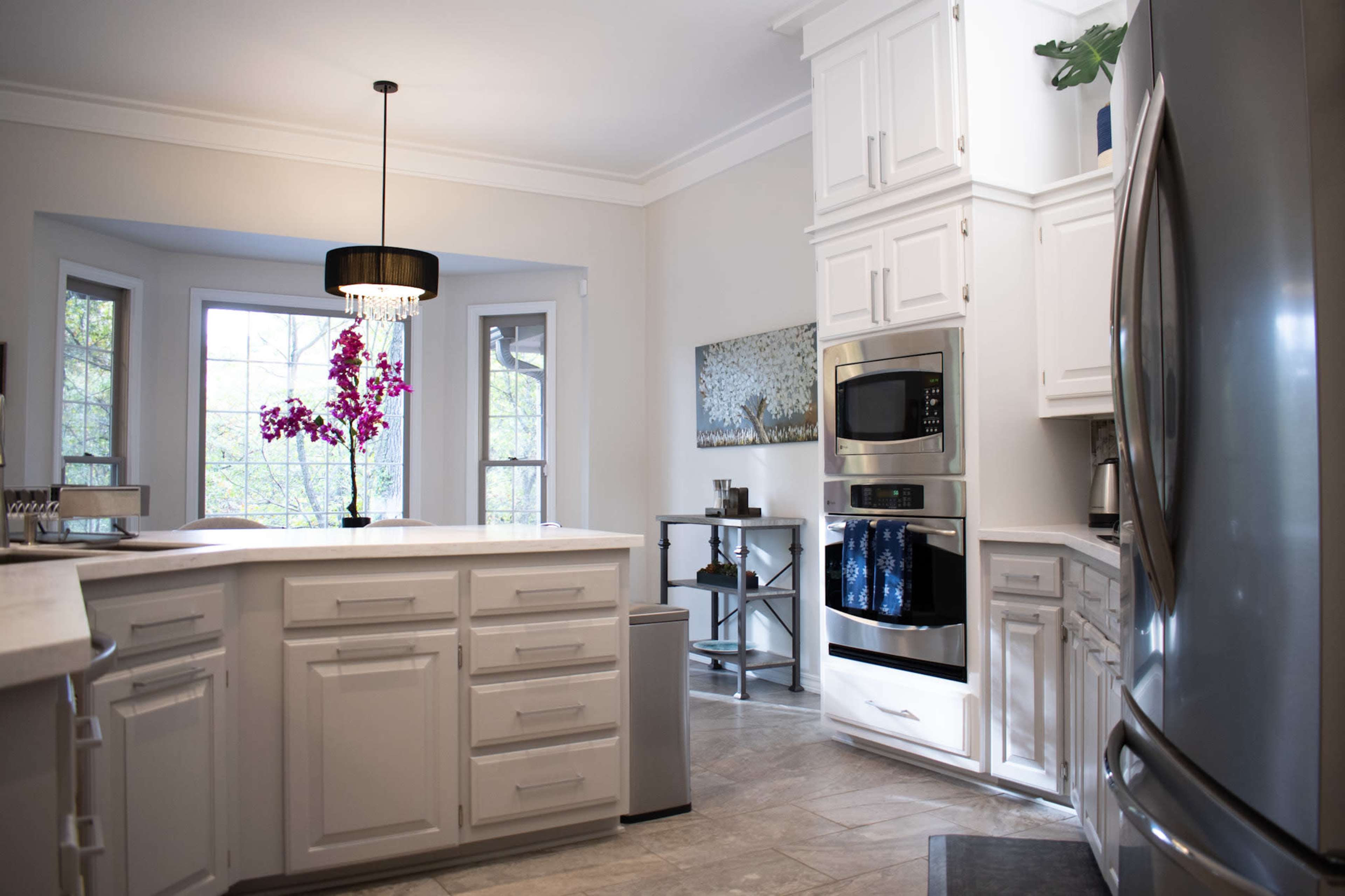 A modern kitchen with white cabinets, a stainless steel refrigerator, and a central island, illuminated by a hanging light fixture above a table with an arrangement of purple flowers.
