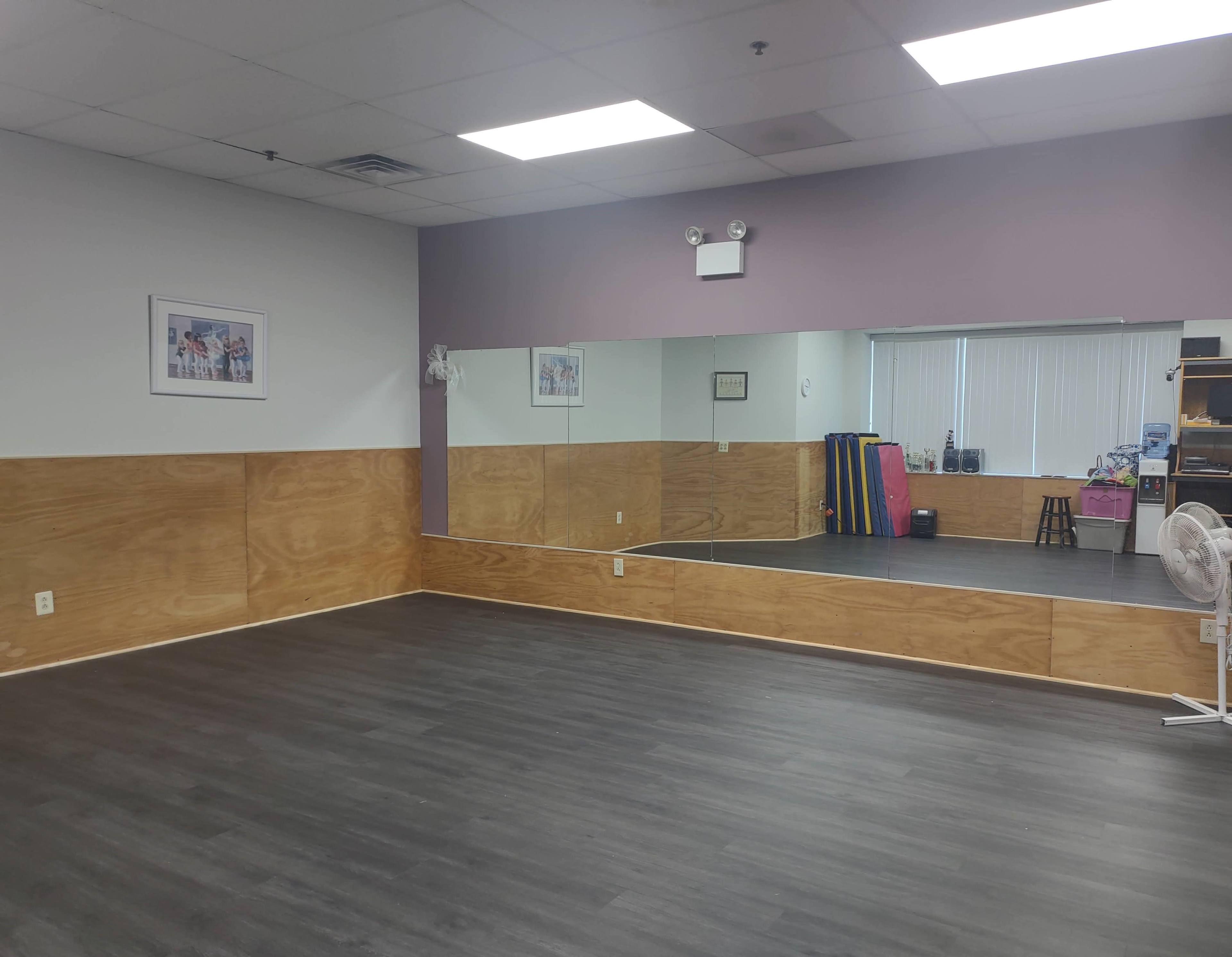 The image shows a spacious, empty room with wooden paneling, a large wall mirror, and exercise equipment stored in the corner.