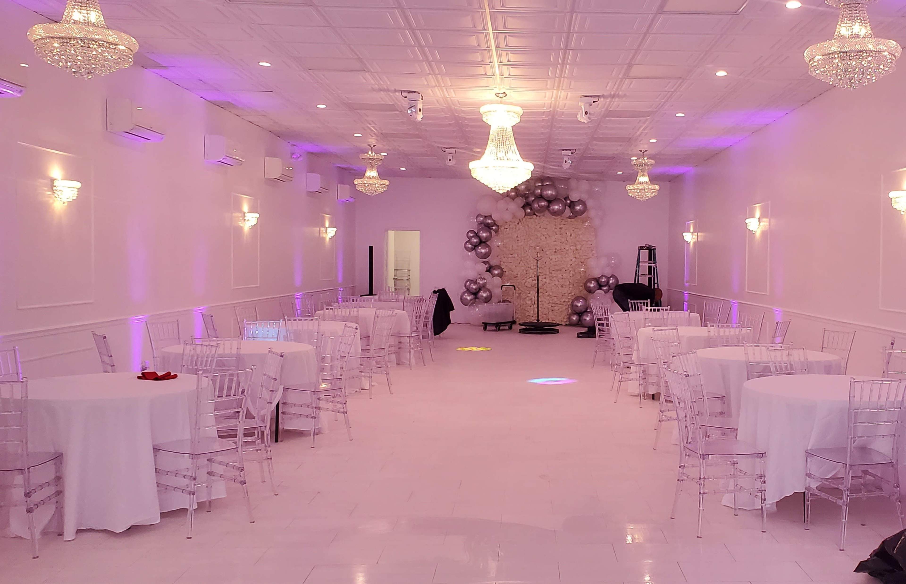 A spacious event hall with white walls, round tables covered in white cloth, and elegant chandeliers, illuminated by soft purple lighting.