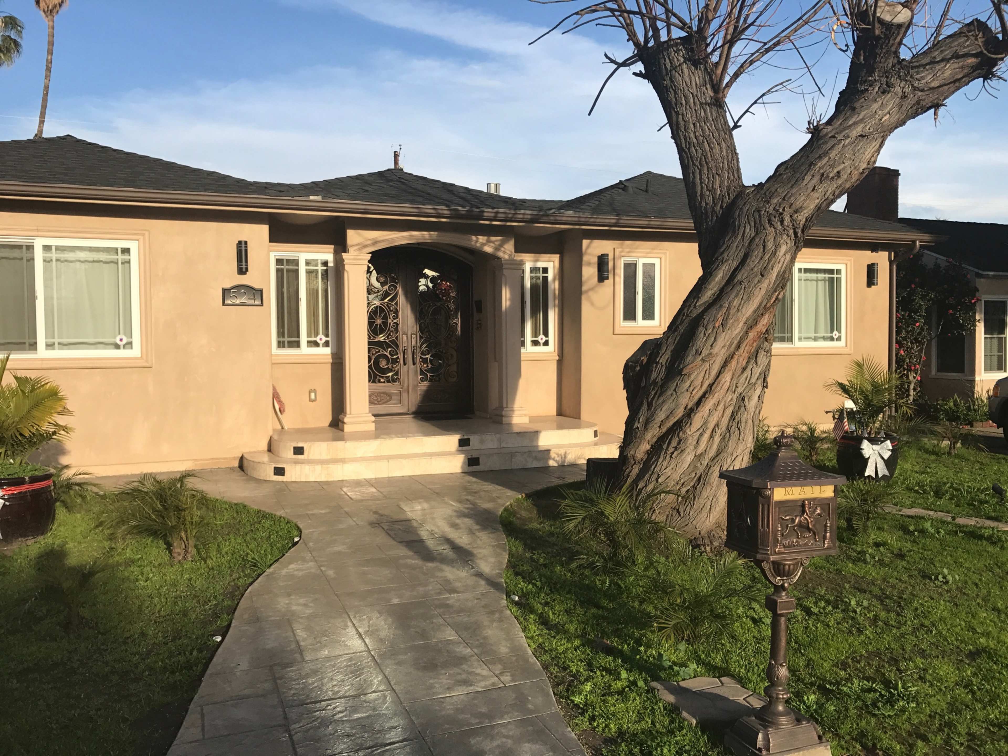 A single-story house with a decorative front door, surrounded by greenery and a large tree, features a concrete walkway leading to the entrance.