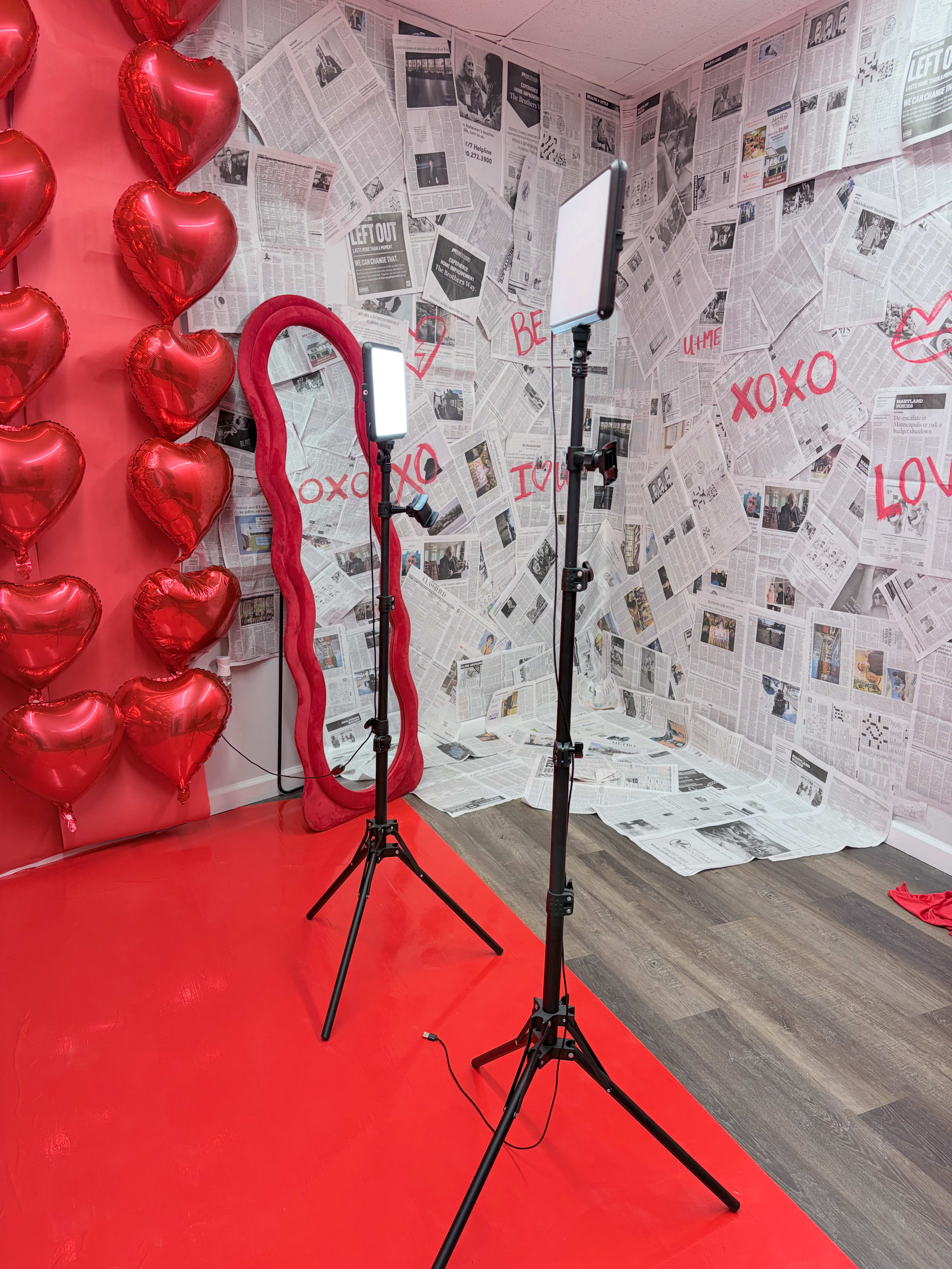 The scene features two light stands with softboxes in front of a red wall adorned with heart-shaped balloons and newspaper clippings.