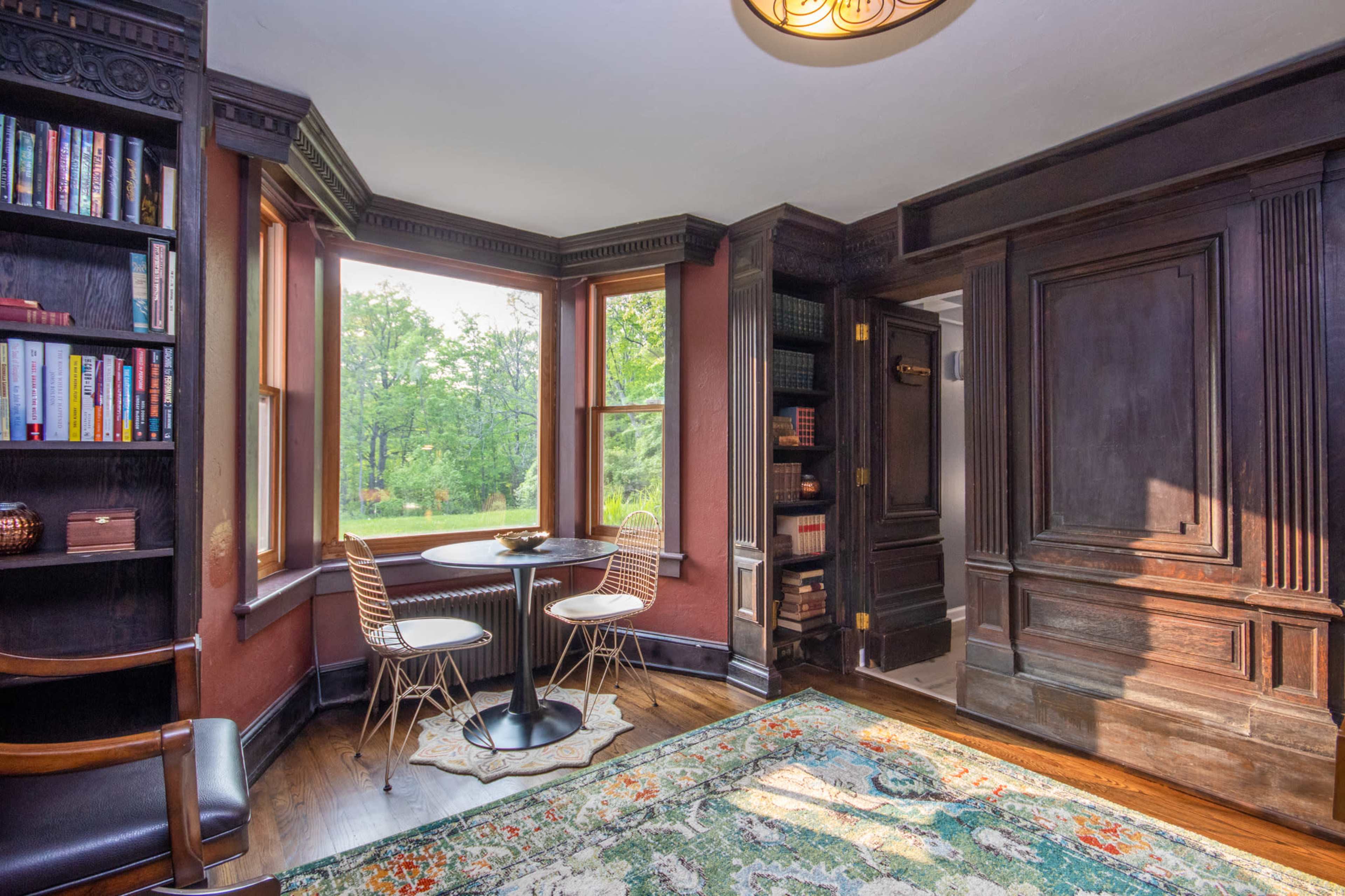 A small dining area with a round table and chairs is positioned near large windows, surrounded by dark wood paneling and bookshelves filled with books.