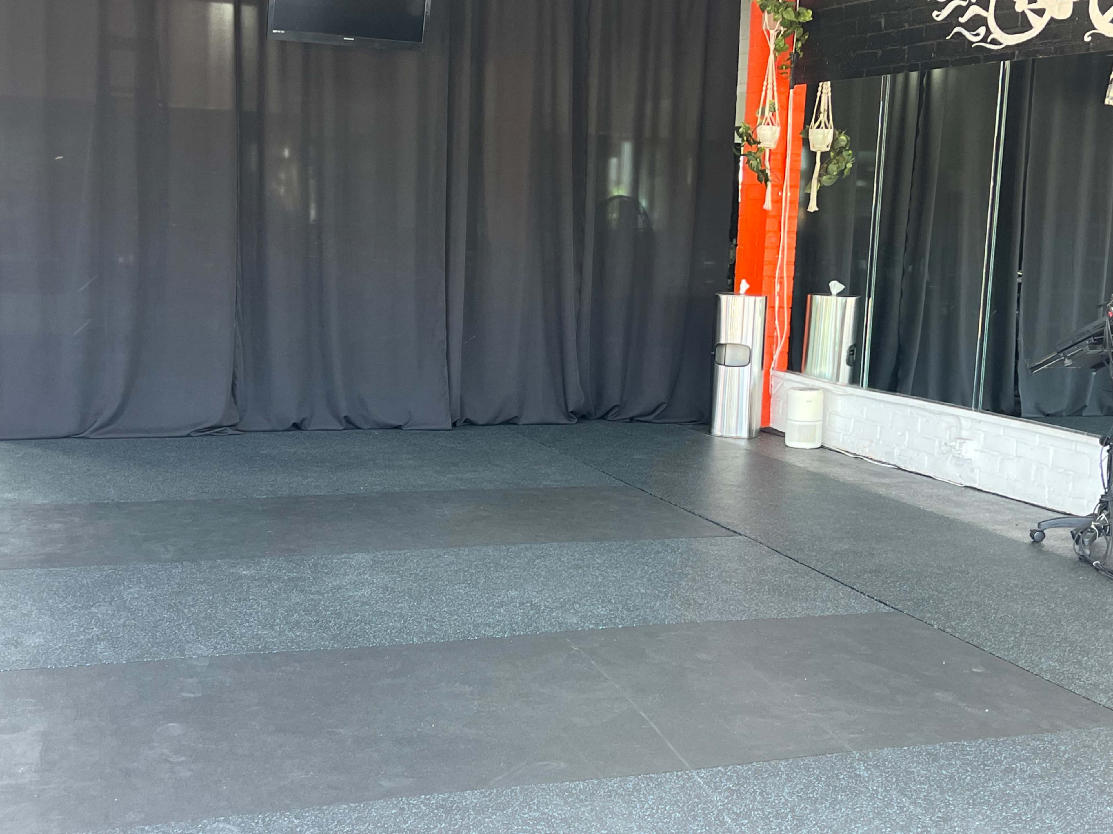 The image shows a spacious studio with black walls, a large mirror, and dark rubber flooring, illuminated by natural light from a window.