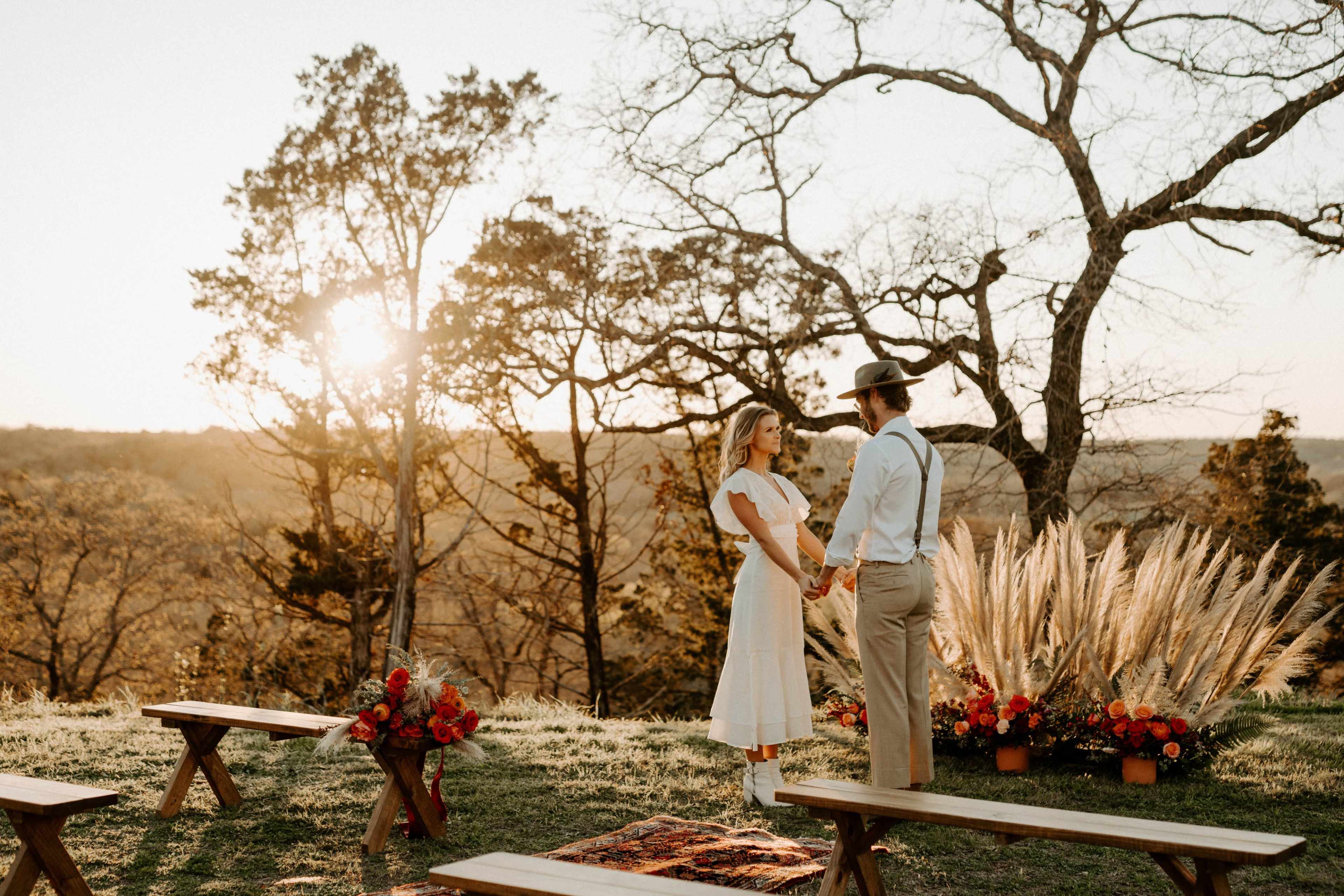 A couple holds hands during a sunset ceremony in an outdoor setting decorated with flowers and wooden benches.