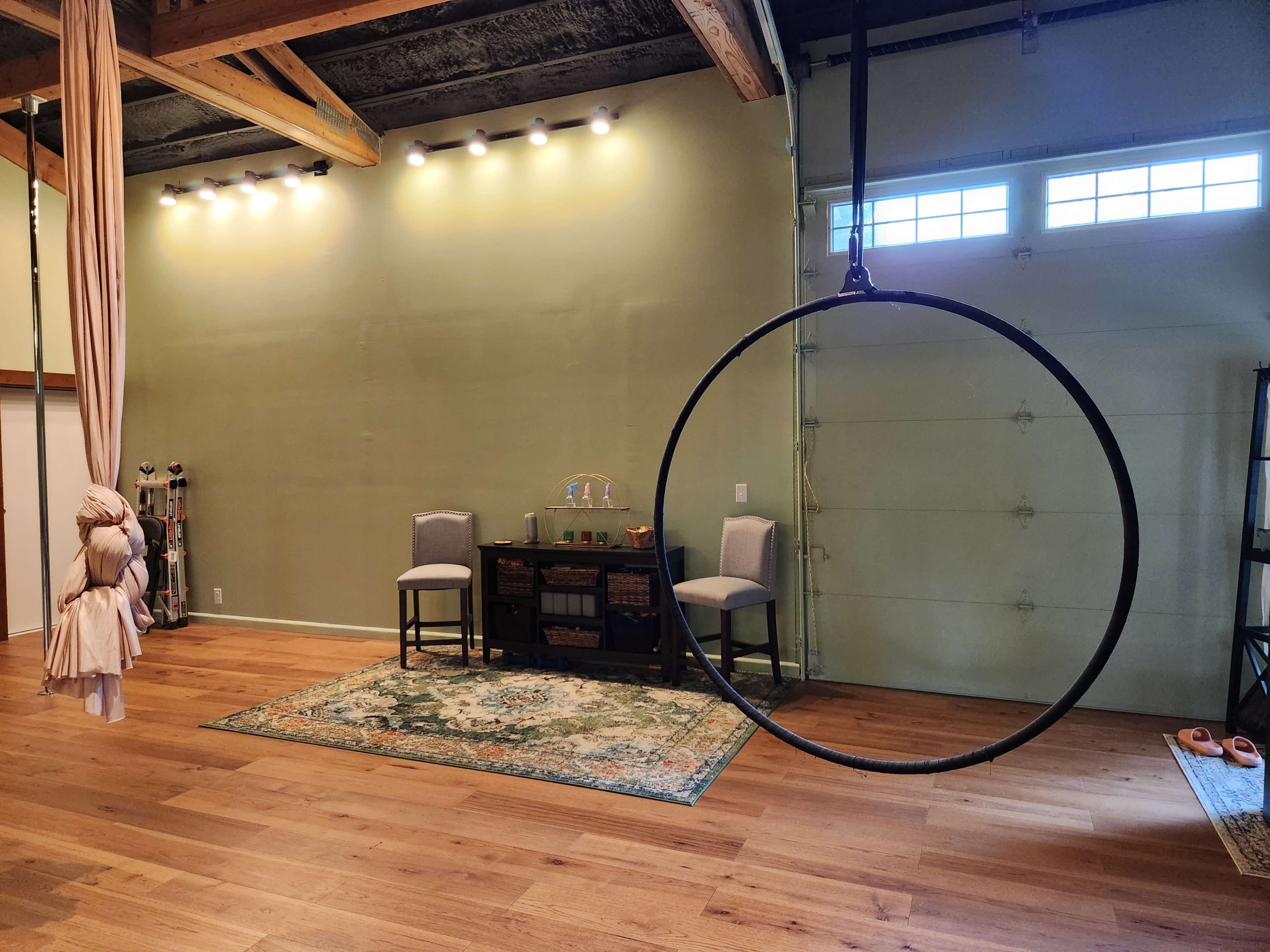 The image shows a spacious room with wooden flooring, featuring a large black aerial hoop suspended from the ceiling, two gray chairs, and a rug on the floor.